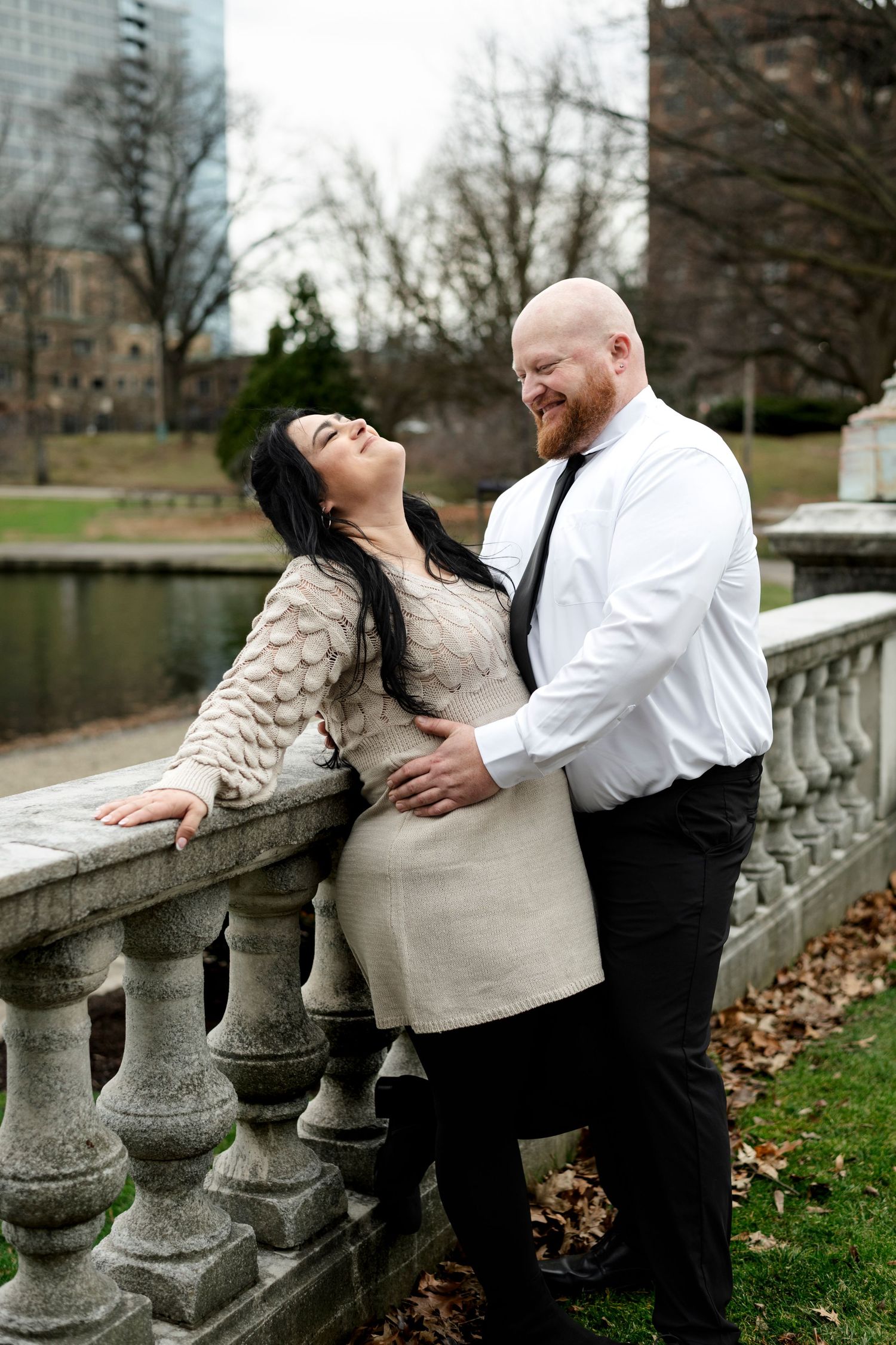 Couple laughing and embracing by stone balustrade in park, woman in beige knit dress, man in white shirt and black tie.