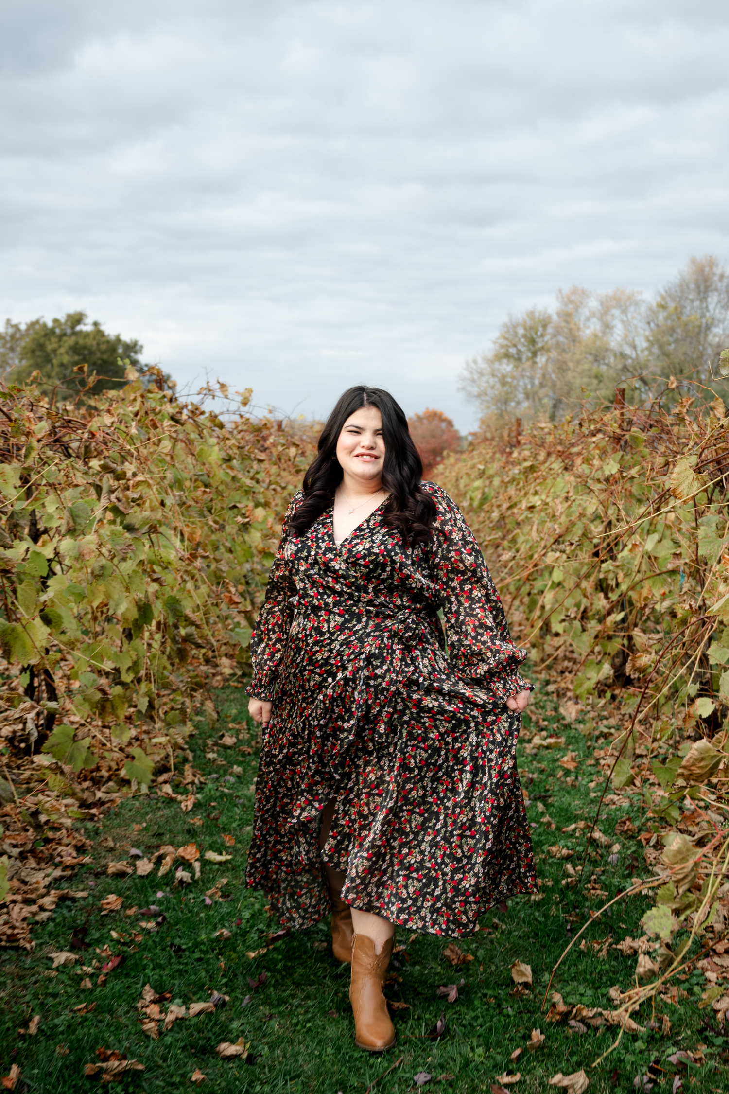 A person in a floral dress poses playfully in a vineyard on a cloudy autumn day.