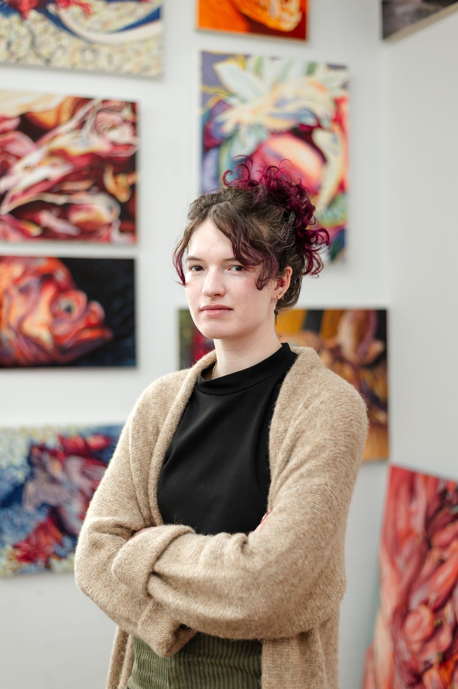 Artist in beige cardigan and black turtleneck stands in front of colorful gallery wall displaying artwork.
