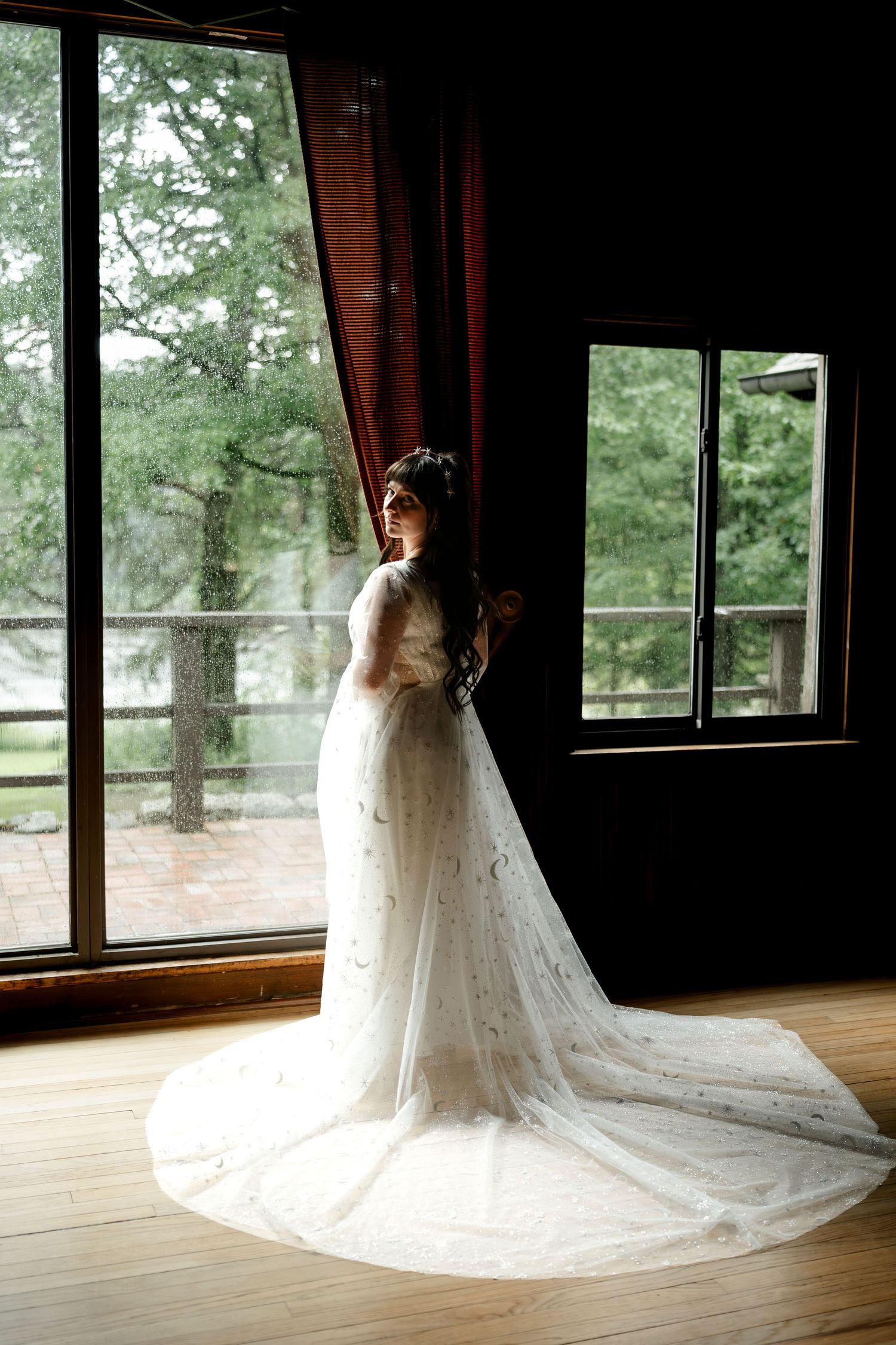 Bride in elegant white lace gown with long train stands by large windows in a rustic wooden venue.