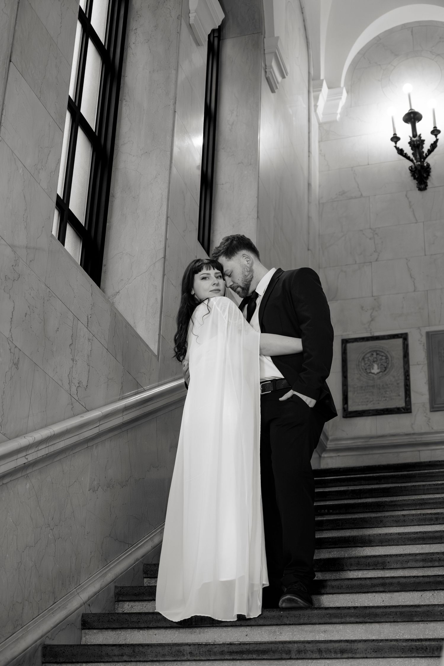 Black and white photo of bride and groom embracing on grand marble staircase in elegant venue.
