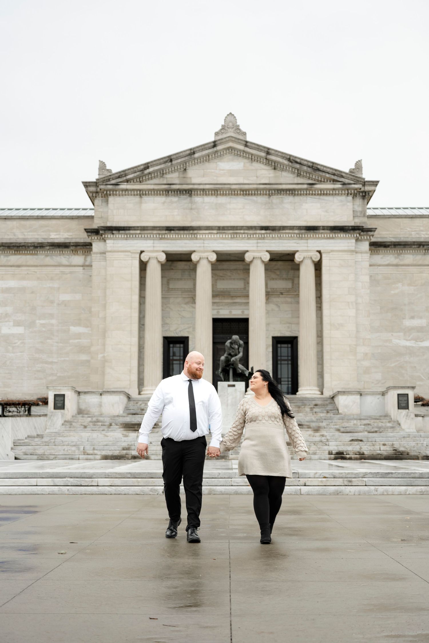 Couple holding hands walking in front of a neoclassical museum building with columns and a bronze statue.