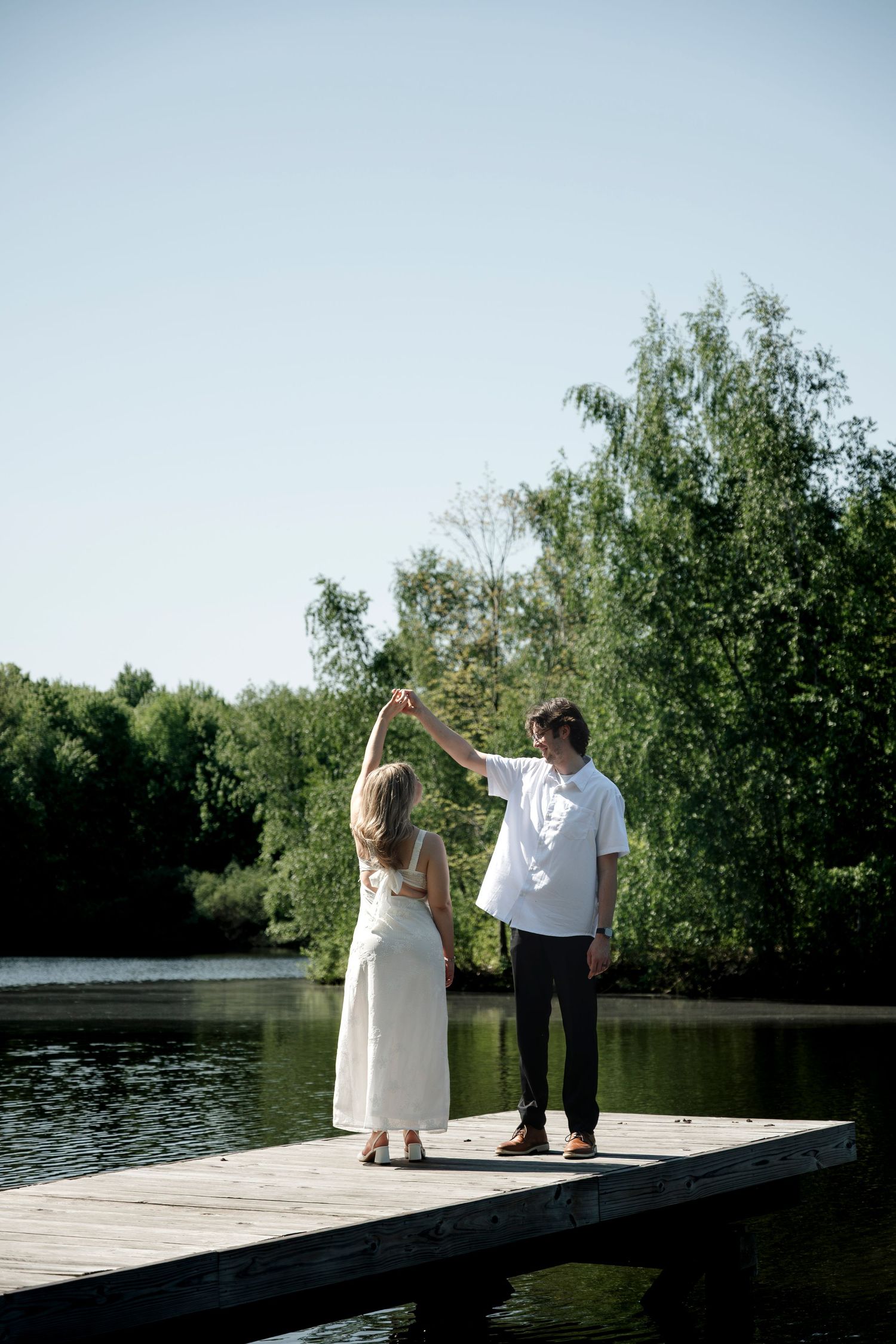 Couple dancing on a wooden dock by a serene lake surrounded by lush green trees on a sunny day.