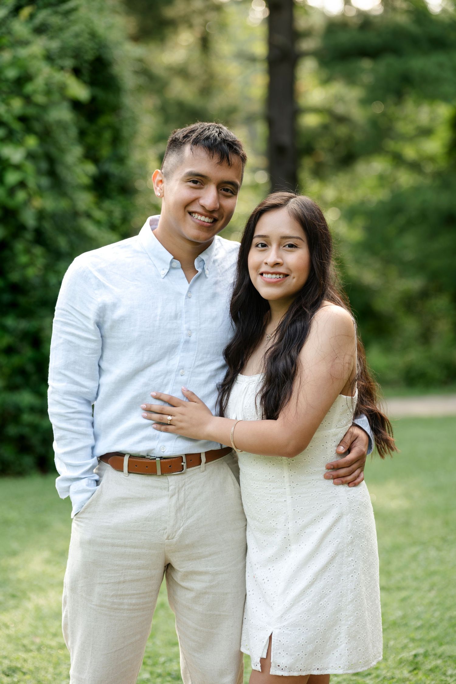 A smiling couple poses together outdoors in a lush green park, wearing coordinated white and light blue outfits.