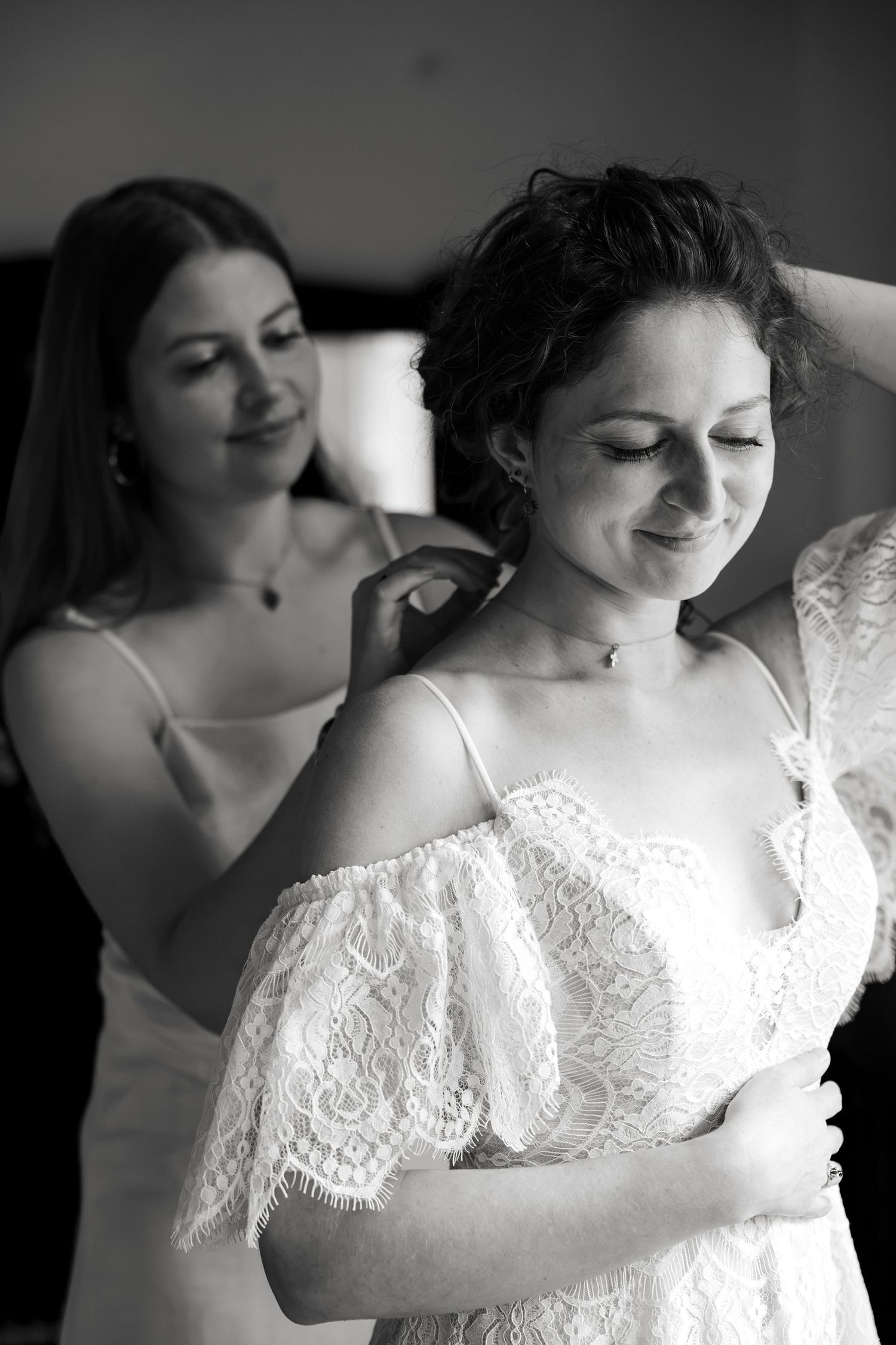 Bridesmaid helping bride into lace wedding dress in black and white photo.