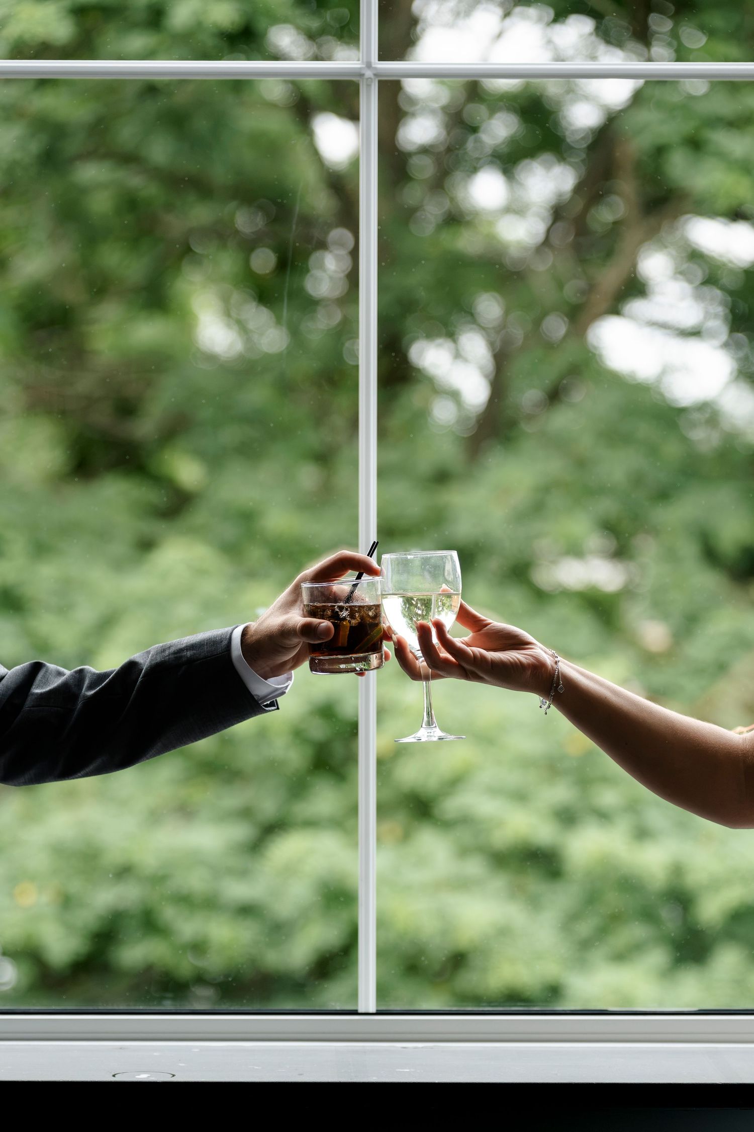Two hands clinking glasses of wine and whiskey in front of a large window with lush green trees.