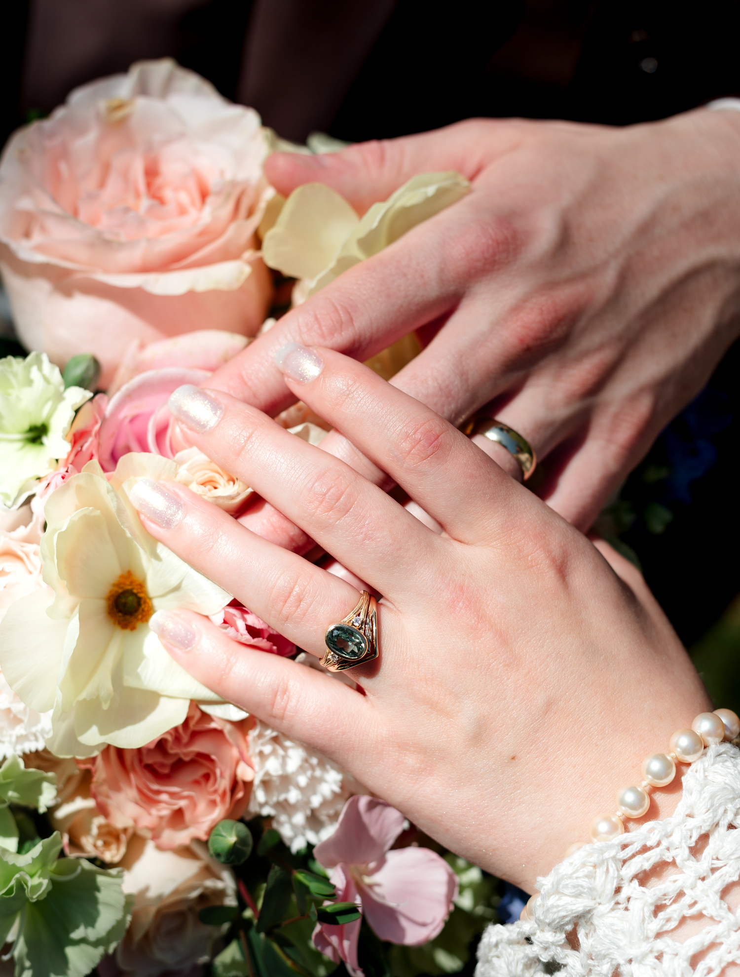 Close up of wedding rings and hands against pink and white floral bouquet.