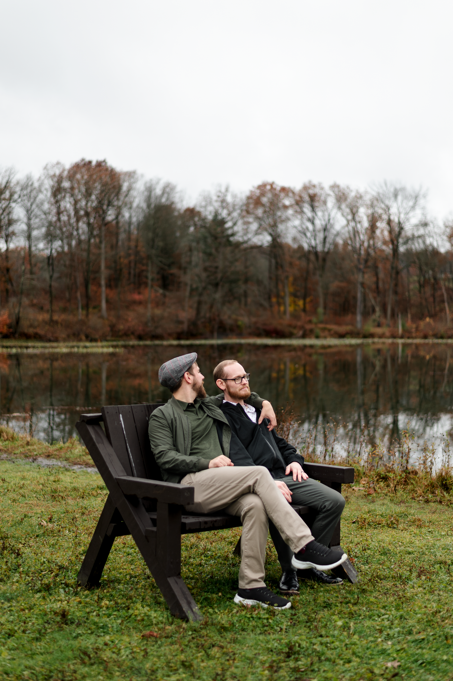 Two men sitting on a dark wooden bench by a serene pond surrounded by autumn trees on an overcast day.