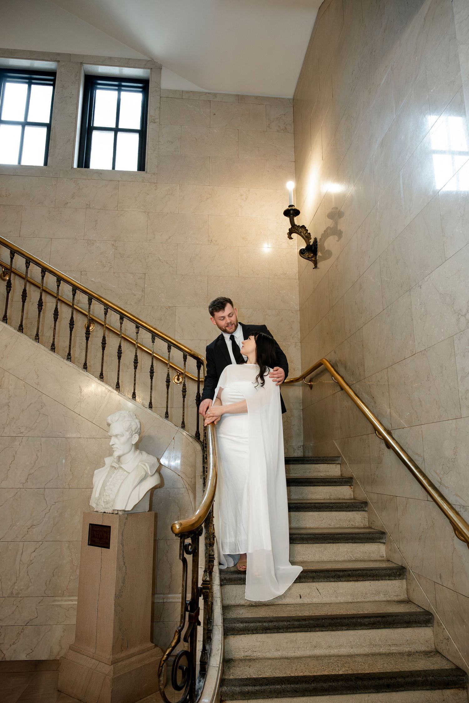 Bride and groom descend marble staircase with gold railing beside classical bust sculpture.