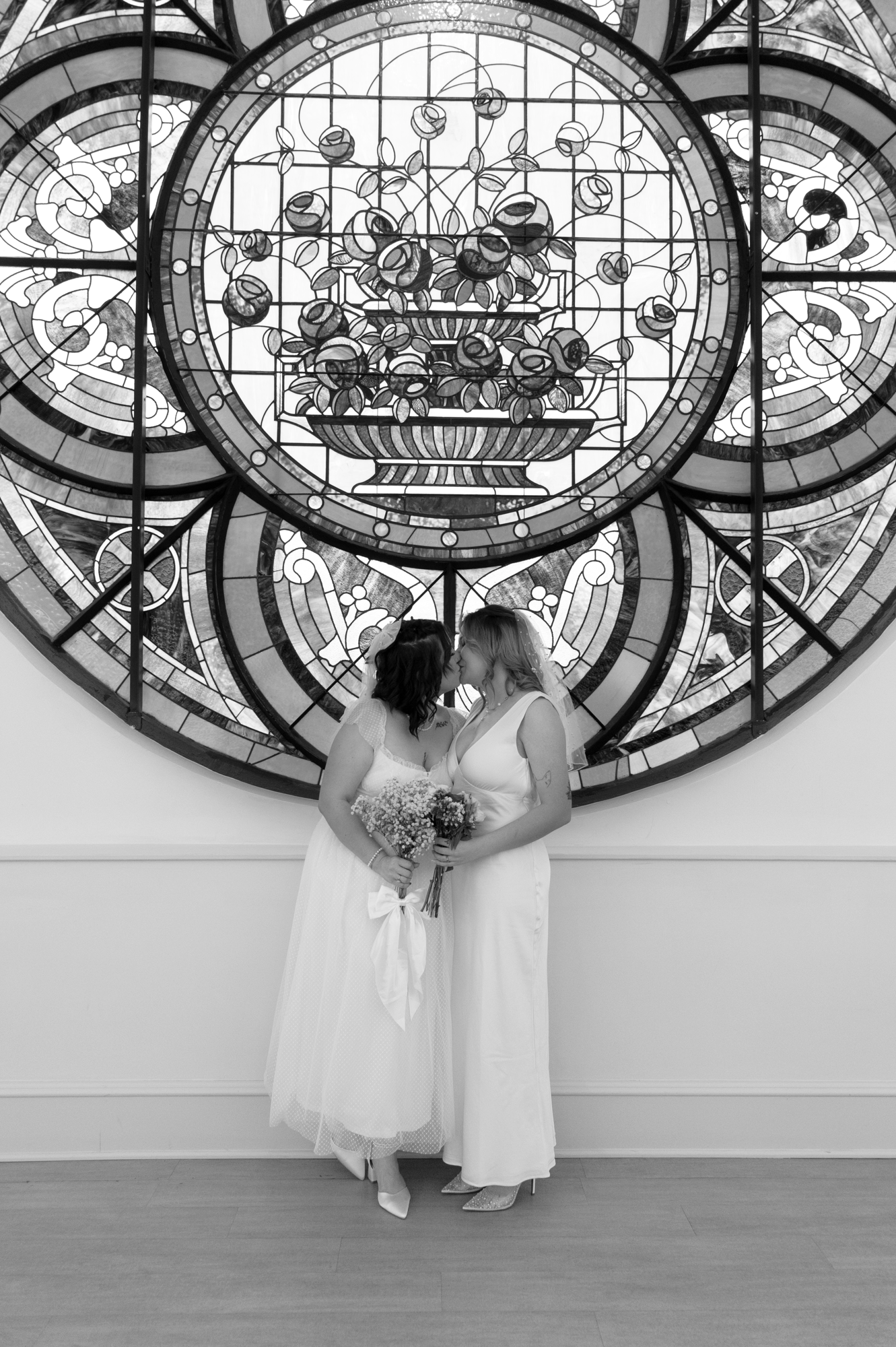 Two brides share a kiss while holding bouquets, standing in front of a large circular stained glass window featuring a floral design.