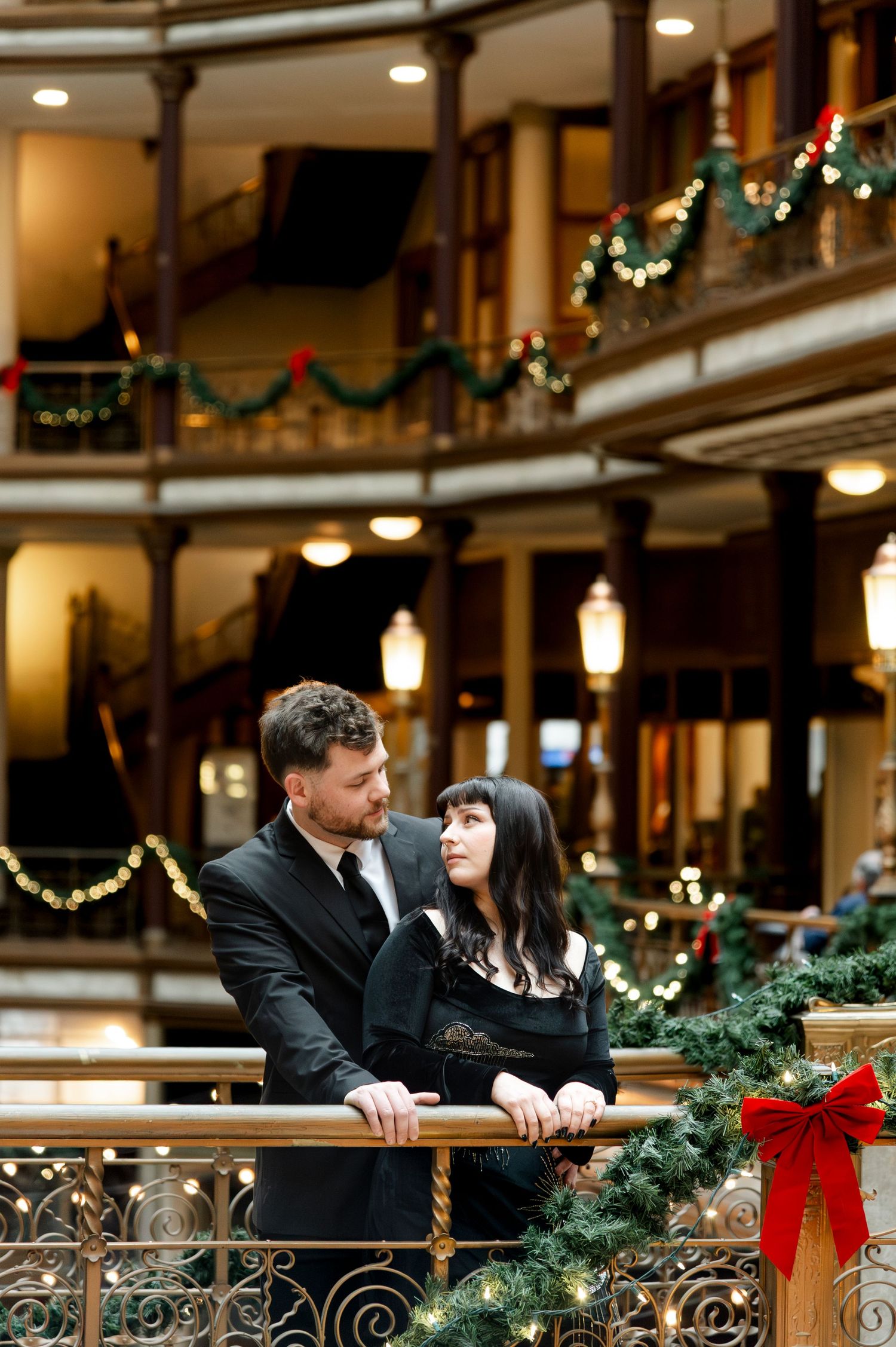 Couple in black attire embracing by festive garland railings in a warmly lit historic building at Christmas.