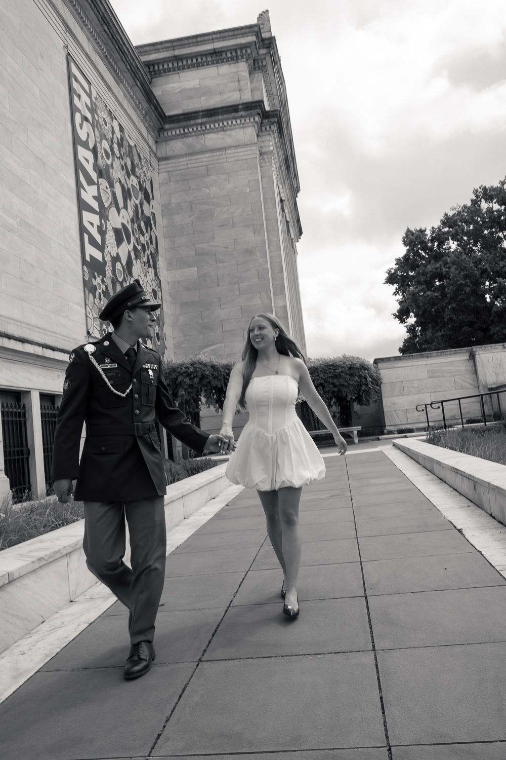 Black and white photo of a couple holding hands, man in military uniform and woman in white dress, walking outside a building.