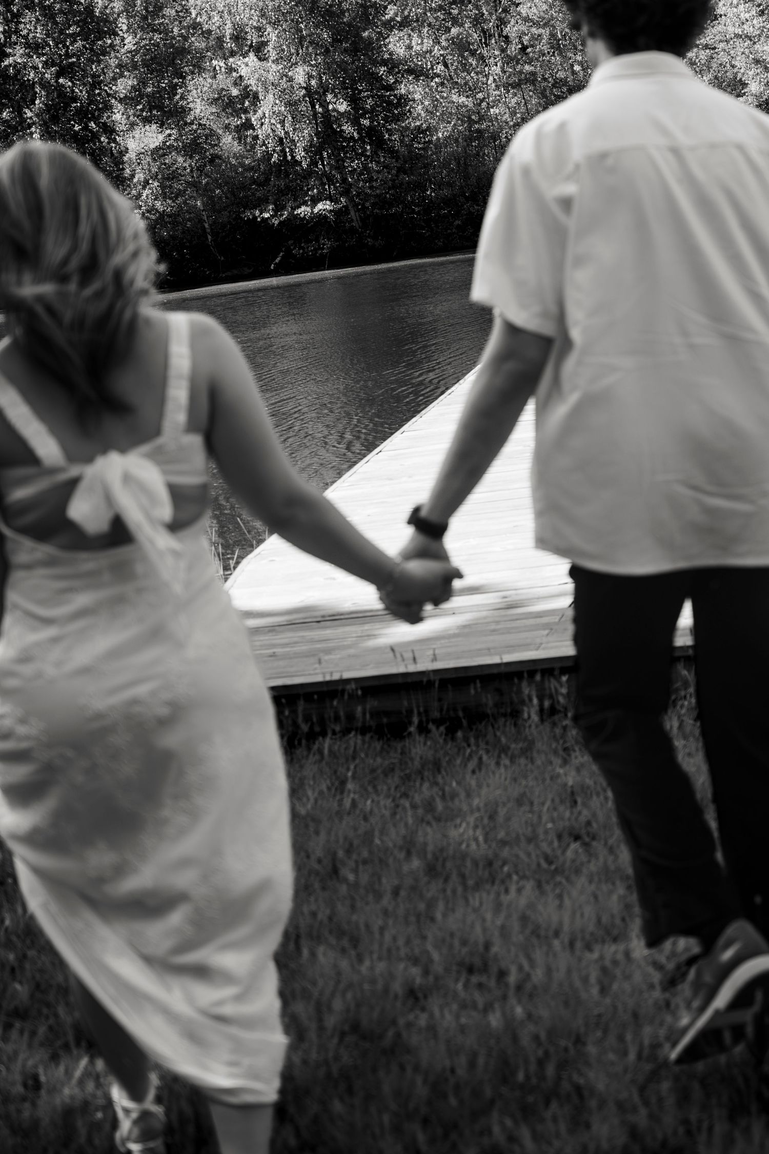 Black and white photo of a couple holding hands walking toward a dock by a serene pond surrounded by trees.
