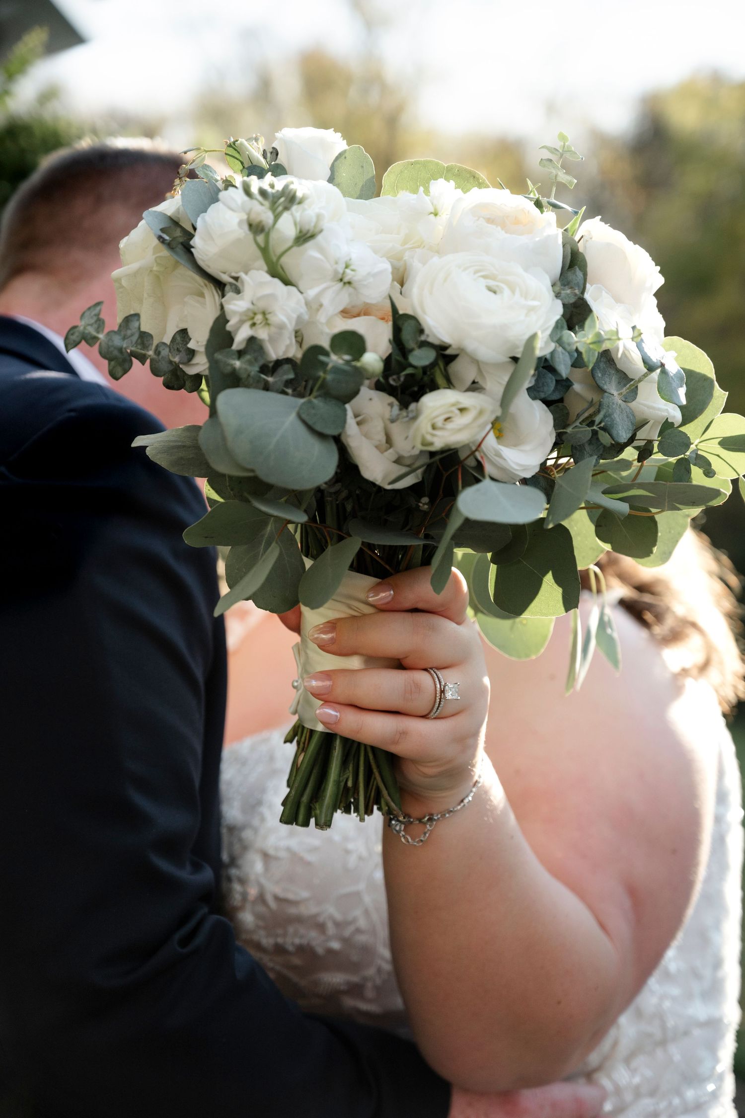 A stunning white wedding bouquet with roses, peonies and eucalyptus leaves held against a white lace dress.