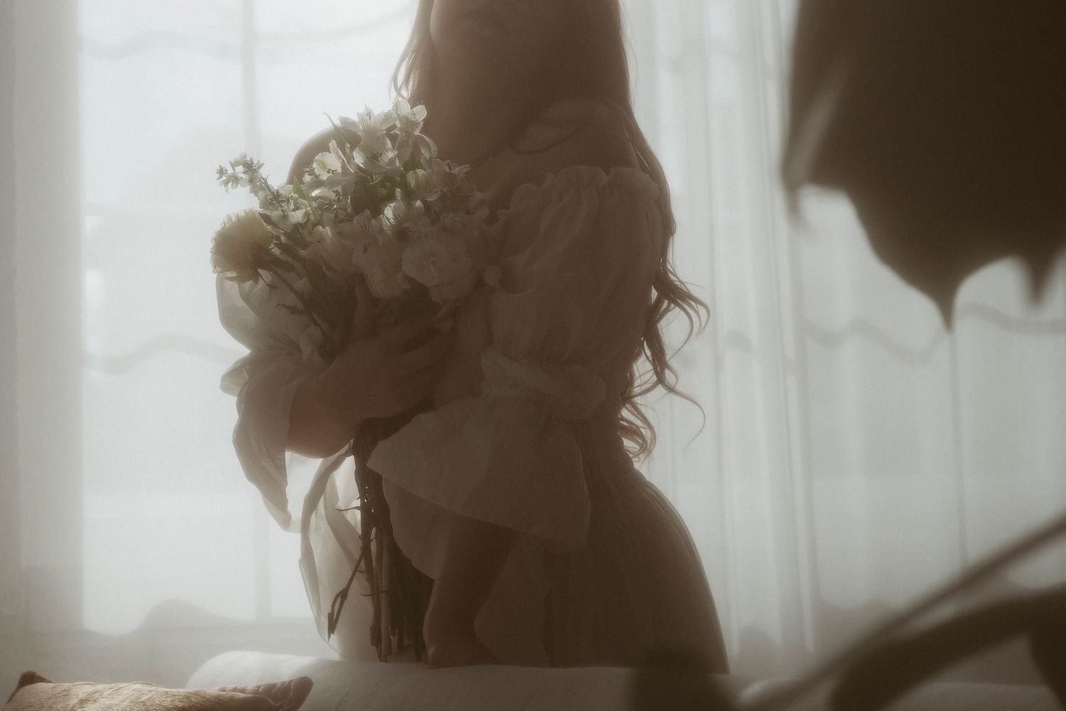 A woman in a white dress holds a delicate bouquet of white flowers near a softly lit sheer curtain window.
