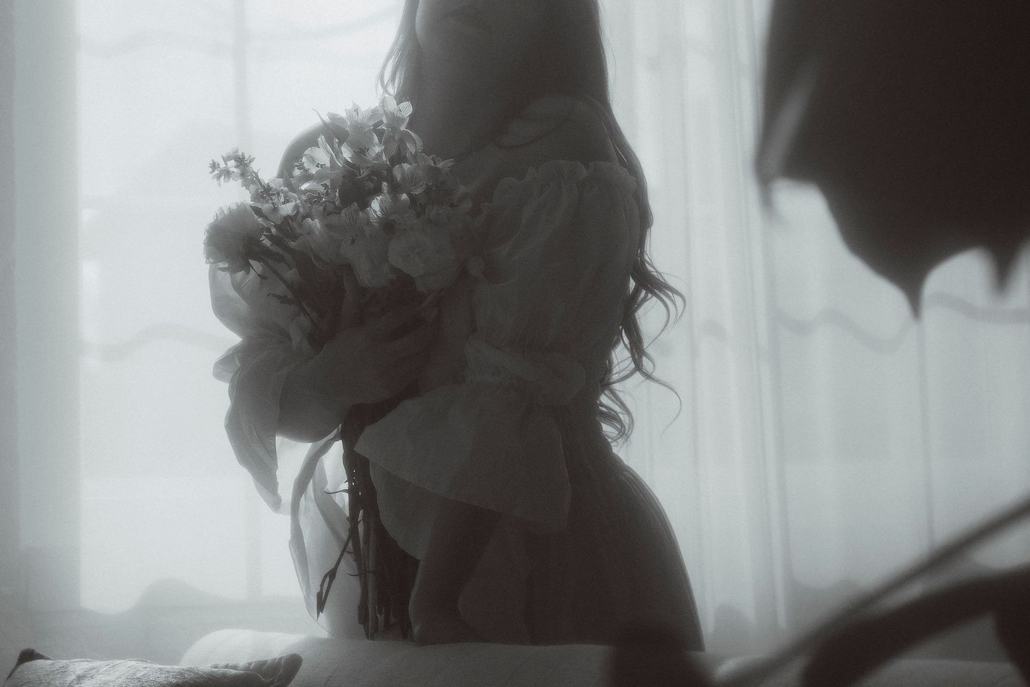 A woman in a soft white dress holds a wildflower bouquet in a dreamy, mist-filled room with sheer curtains.