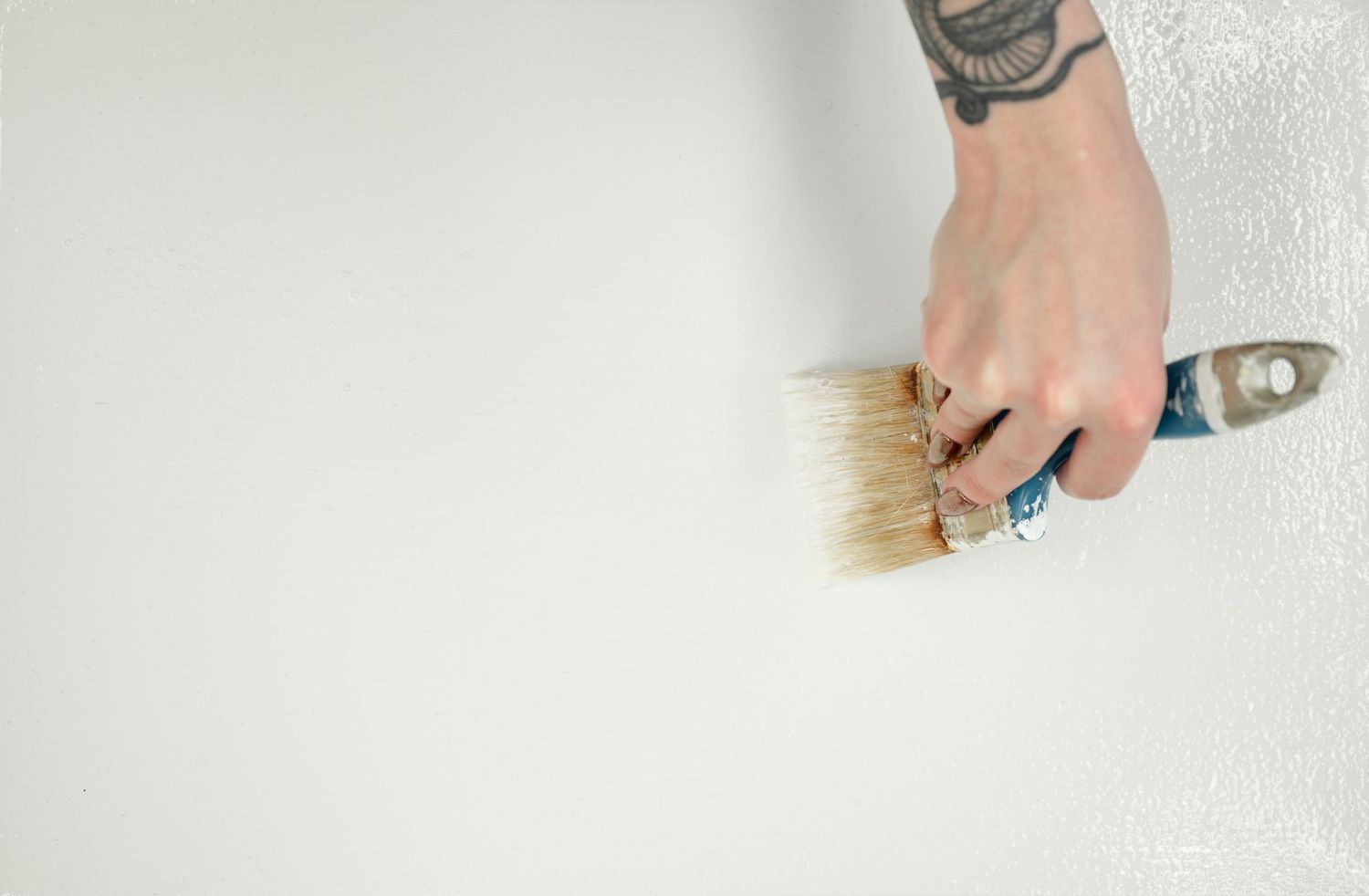 A tattooed hand holds a paintbrush against a white wall while painting.