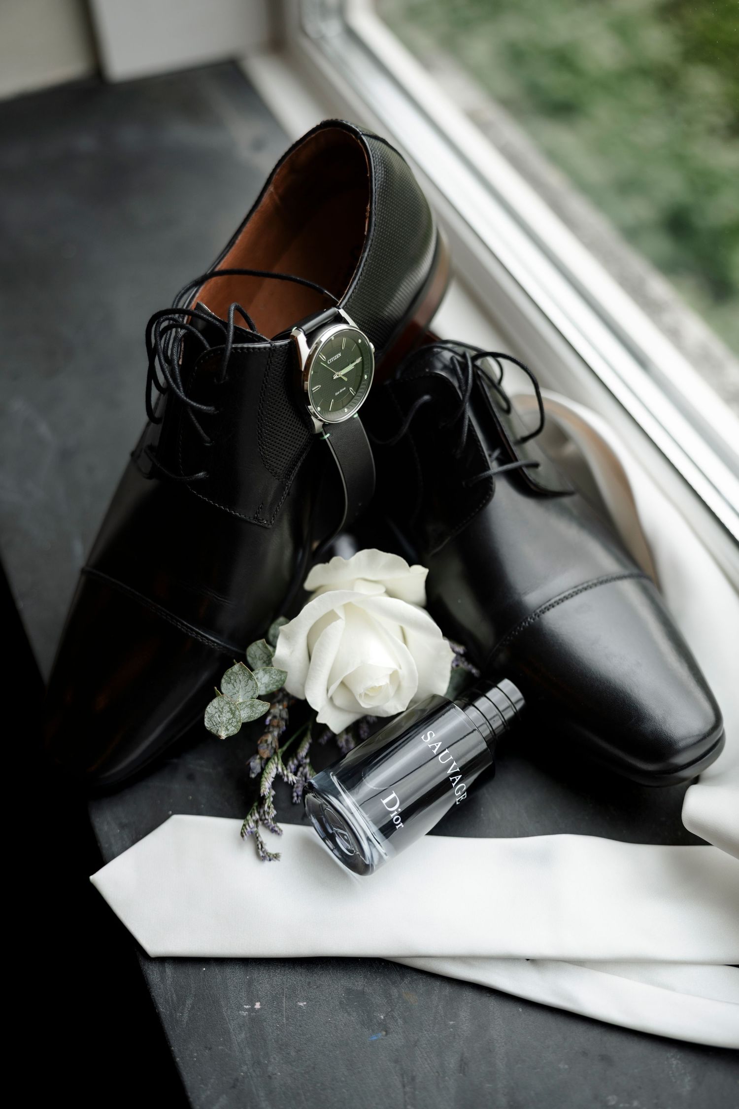 Black dress shoes with a watch, white rose boutonniere, cologne, and white pocket square arranged for a wedding day.