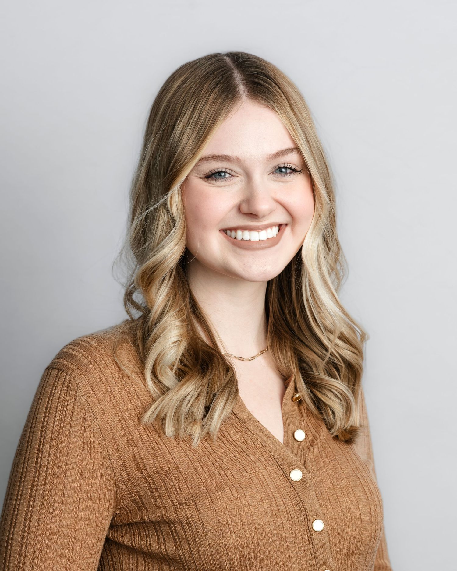 A person wearing a brown sweater smiles warmly while posing against a light gray background in a professional headshot series.
