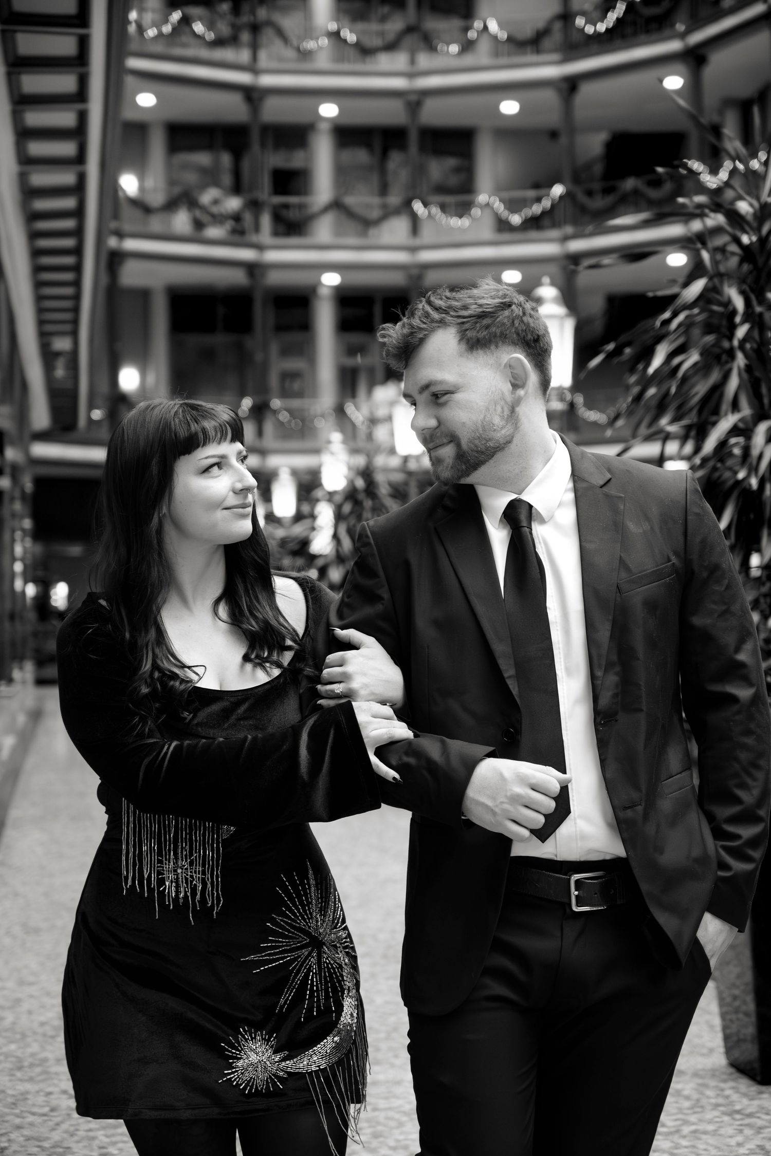 Black and white photo of an elegant couple in formal attire sharing a romantic moment in a grand hotel lobby.