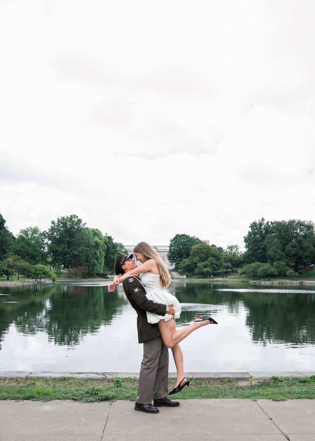 Couple sharing romantic kiss by a serene lake, groom lifting bride in white dress outdoors.