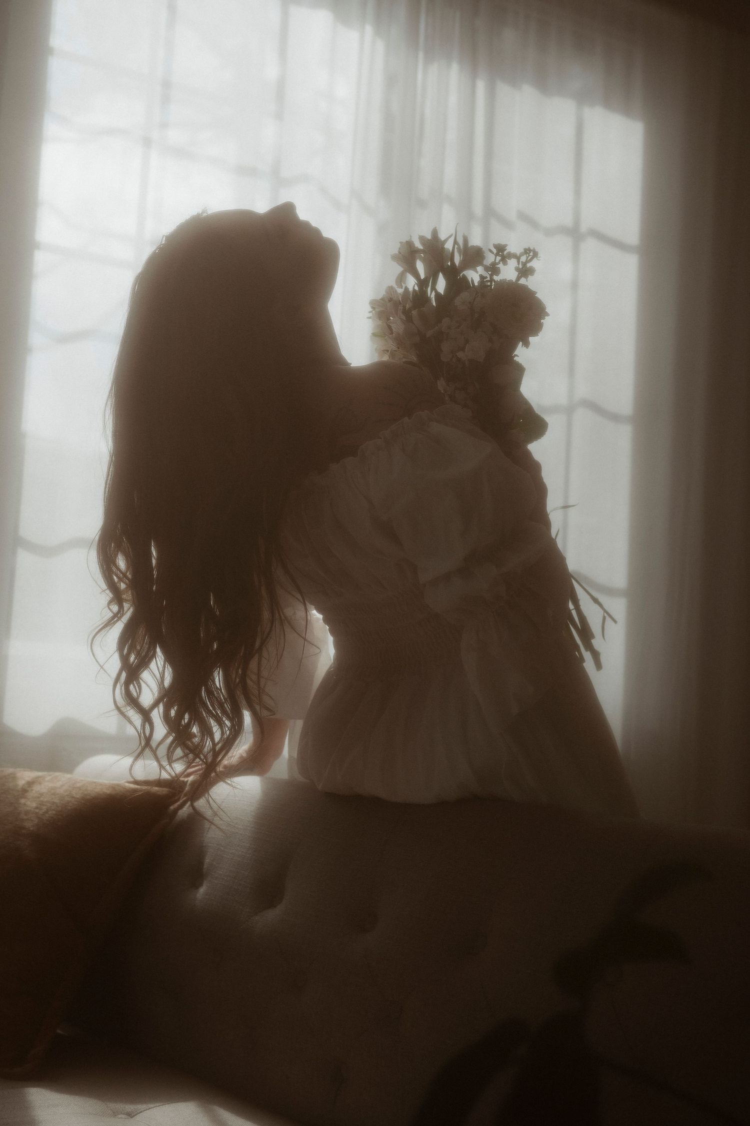 Silhouette of woman in white dress holding flowers by a softly lit window.