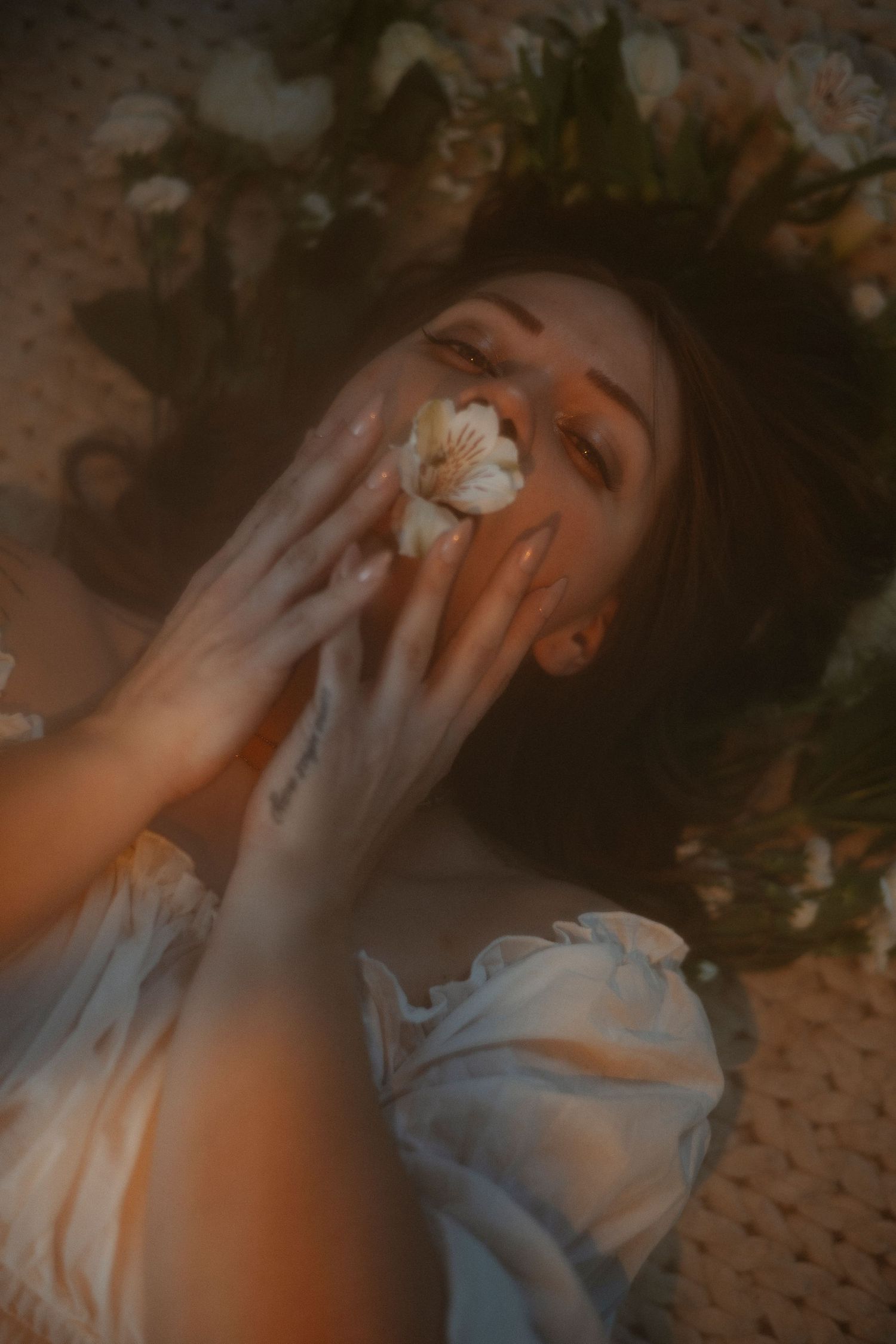 Woman lying among flowers holding a white bloom near her face in warm golden light.