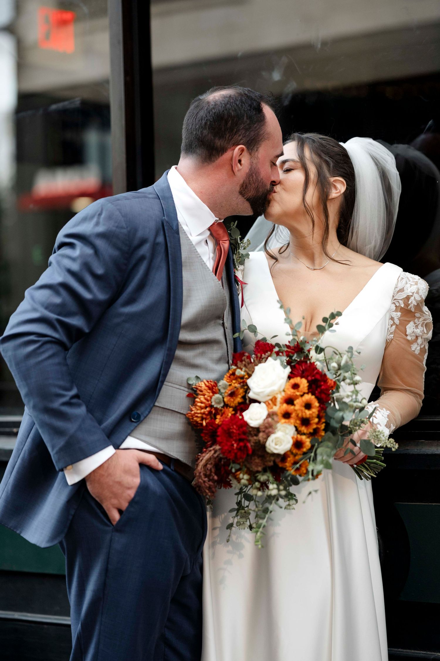 A wedding couple shares a romantic kiss while holding a vibrant orange and red autumn bouquet.