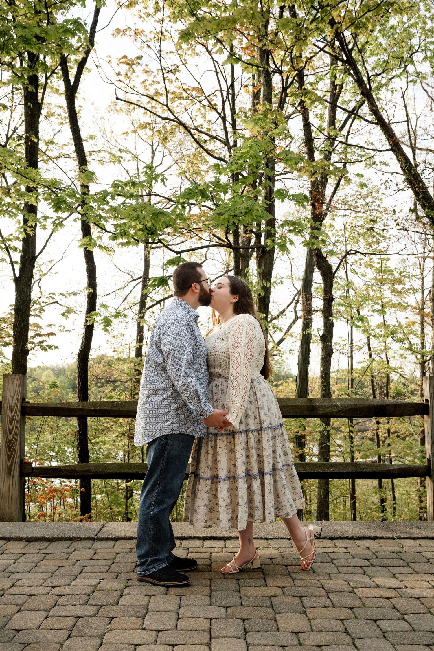 Couple sharing a romantic kiss on a brick patio surrounded by lush green trees in an outdoor woodland setting.