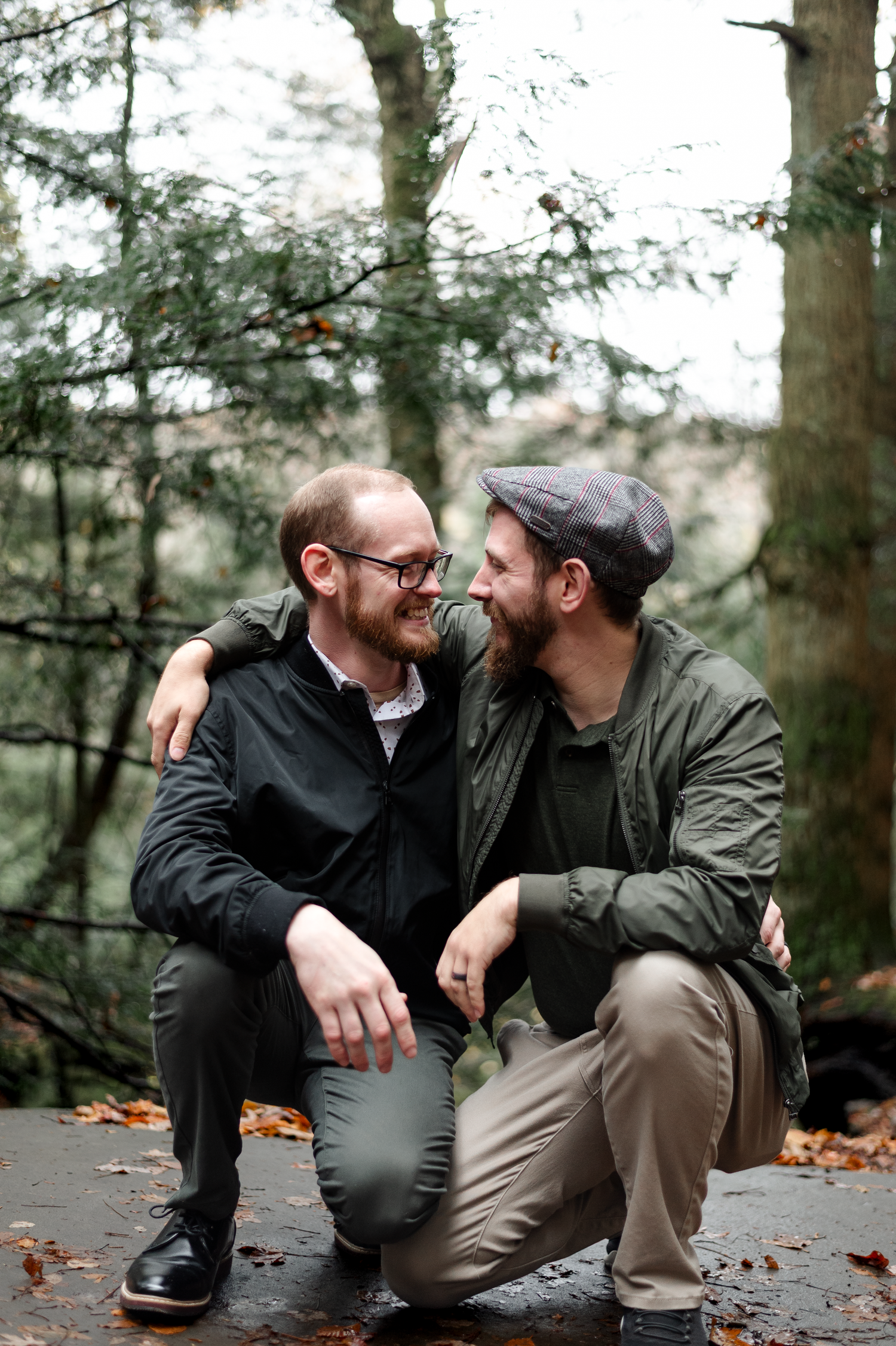 Two men smiling at each other in a forest, one with glasses and one wearing a cap, sharing a warm embrace outdoors.