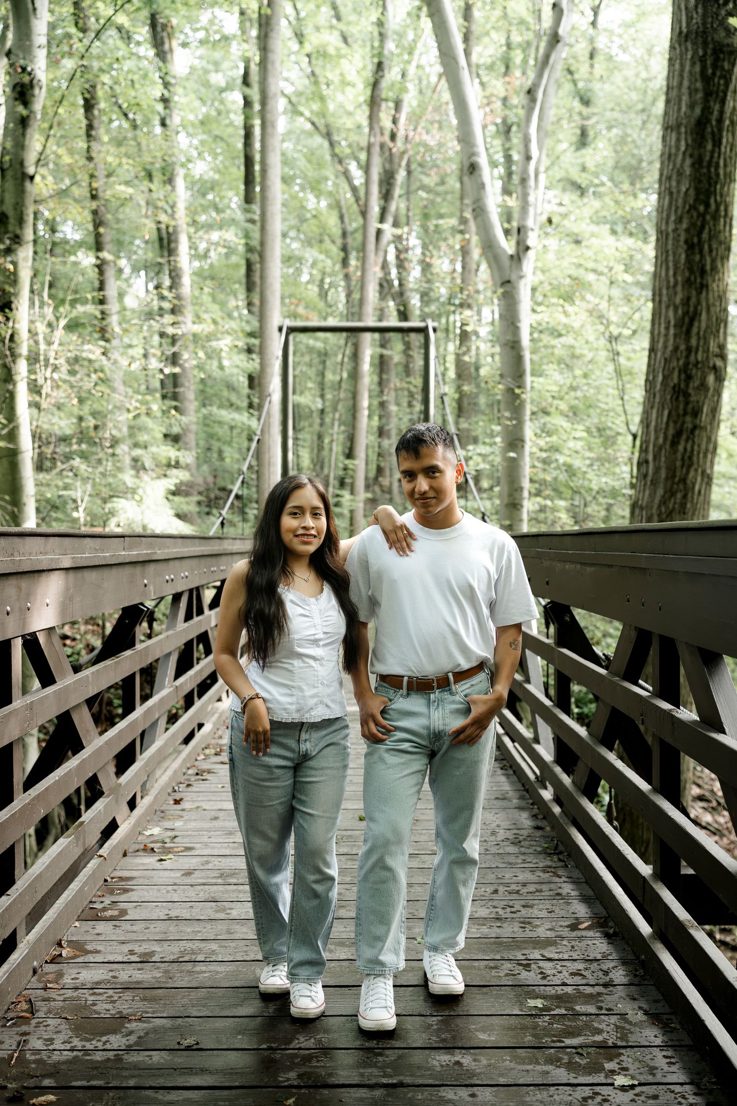 Couple in matching white tees and jeans posing on a wooden forest bridge surrounded by tall trees.