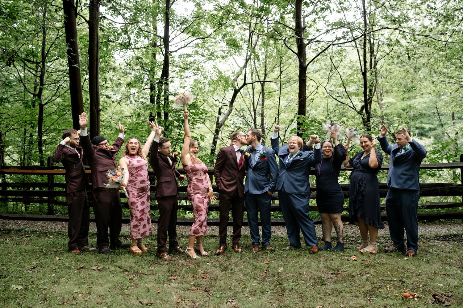 Wedding party celebrating outdoors in forest setting, wearing maroon and navy attire, raising arms joyfully near wooden fence.