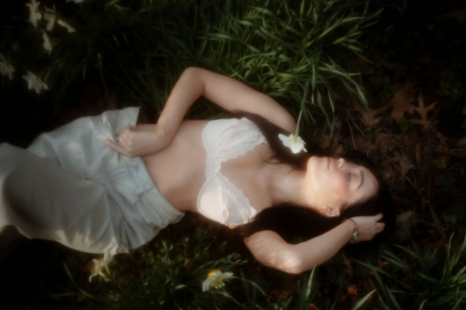 A woman in a white dress lies peacefully among green grass and white flowers in soft, dreamy lighting.