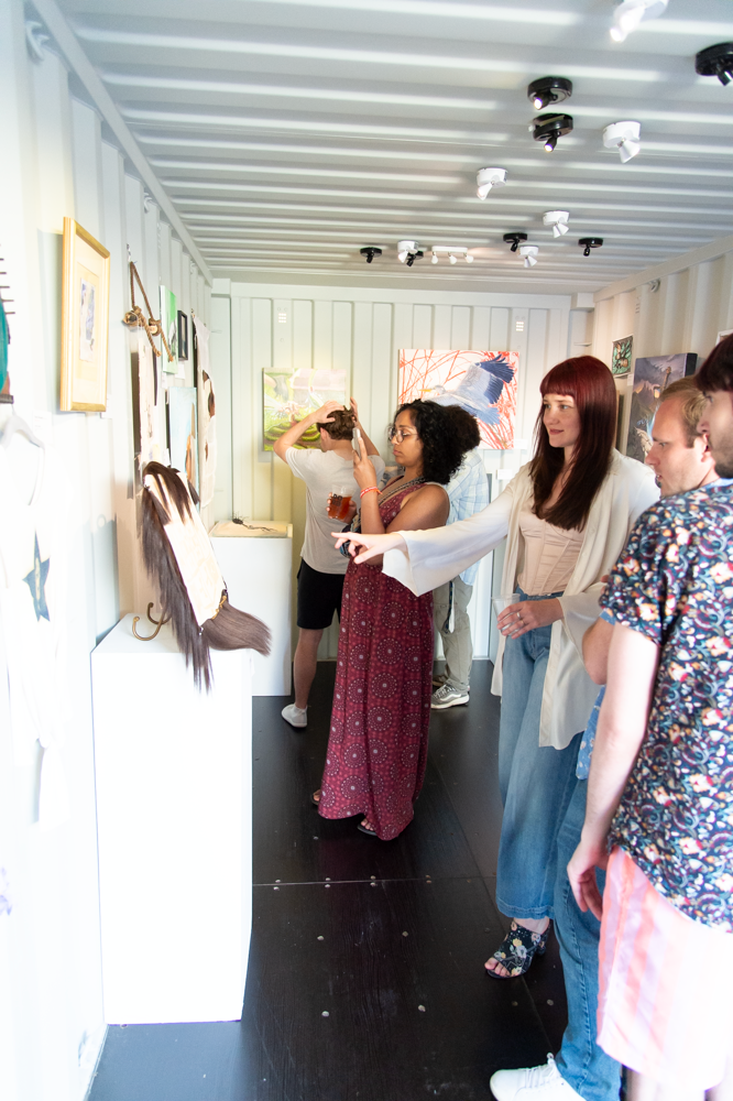 A group of visitors viewing artwork displayed on a white wall in a narrow gallery space with exposed beam ceiling.