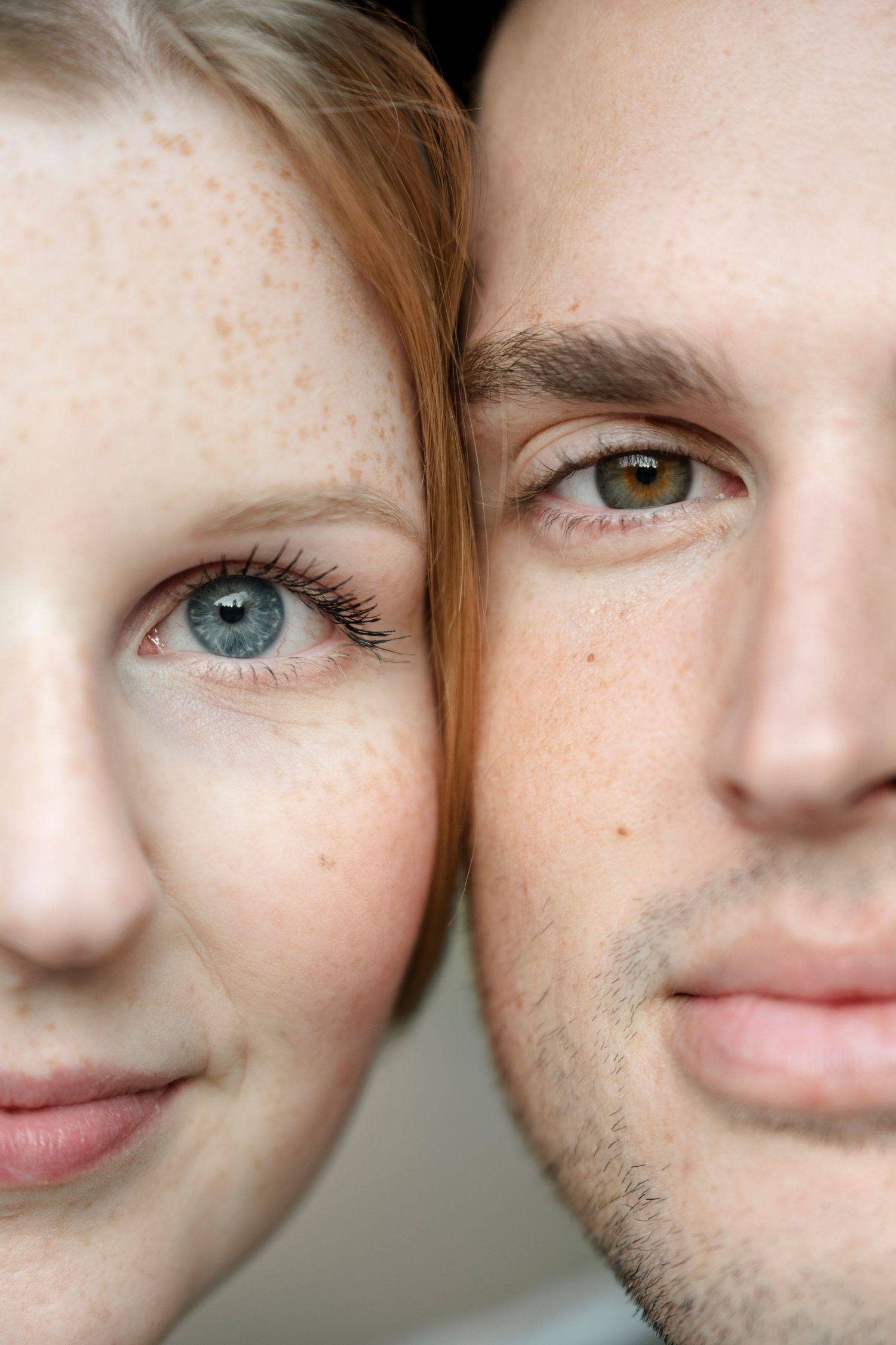 Close-up portrait of a freckled redhead woman with blue eyes and a brown-eyed man with stubble, faces side by side.