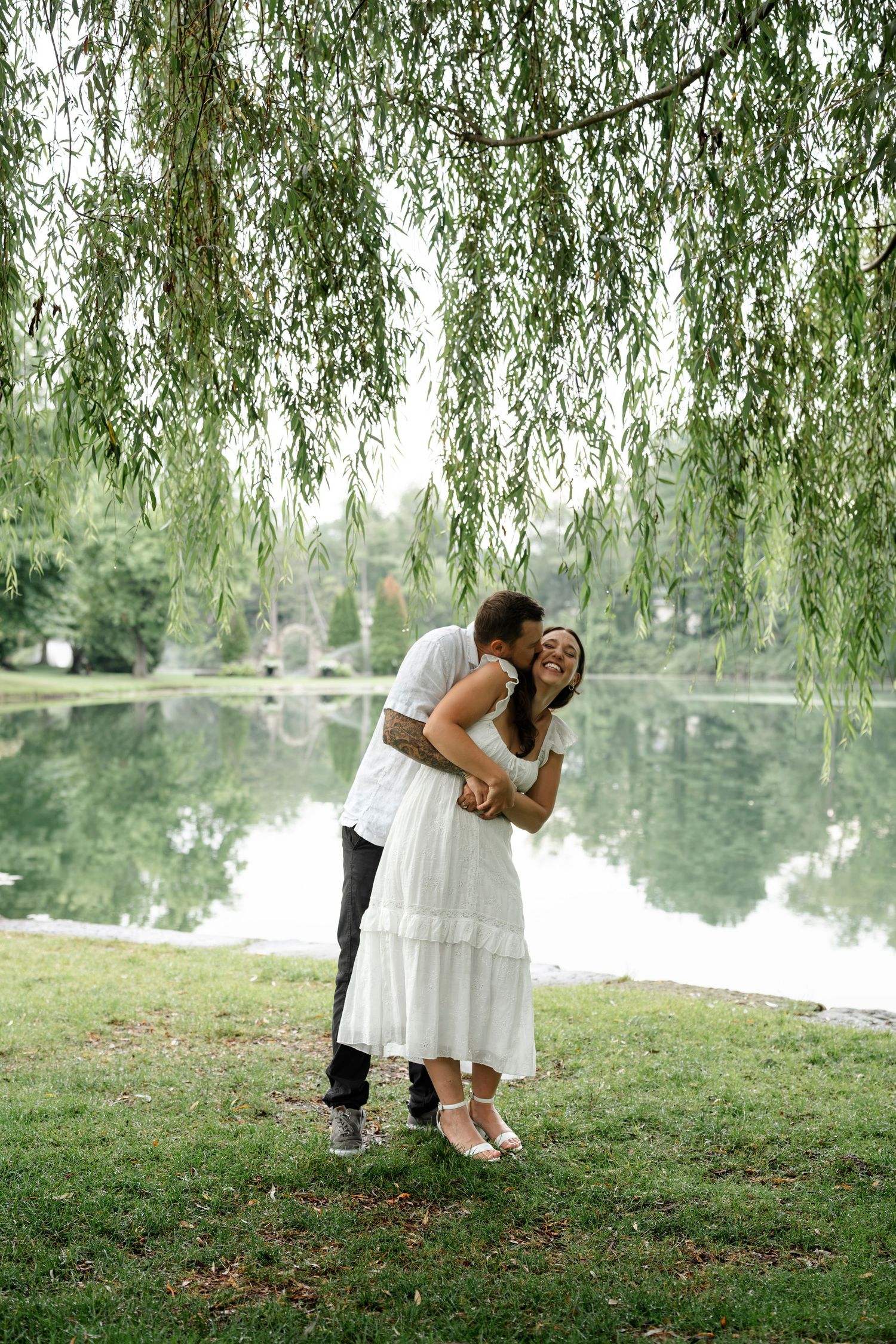 Couple embracing and kissing under a weeping willow tree by a serene pond in a lush green park.