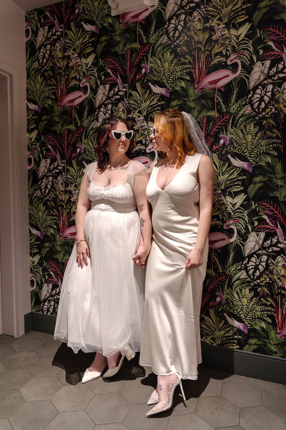 Two people in white dresses pose against a dark floral wallpaper background.