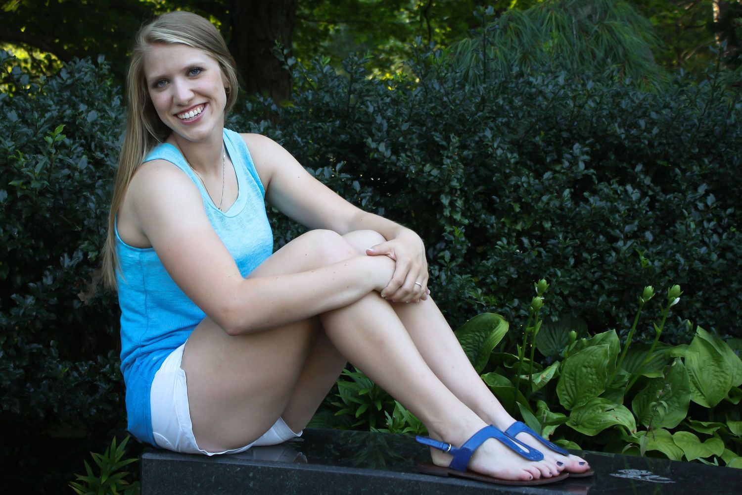 Someone sits on a rock in a garden wearing a blue tank top and shorts on a sunny summer day.