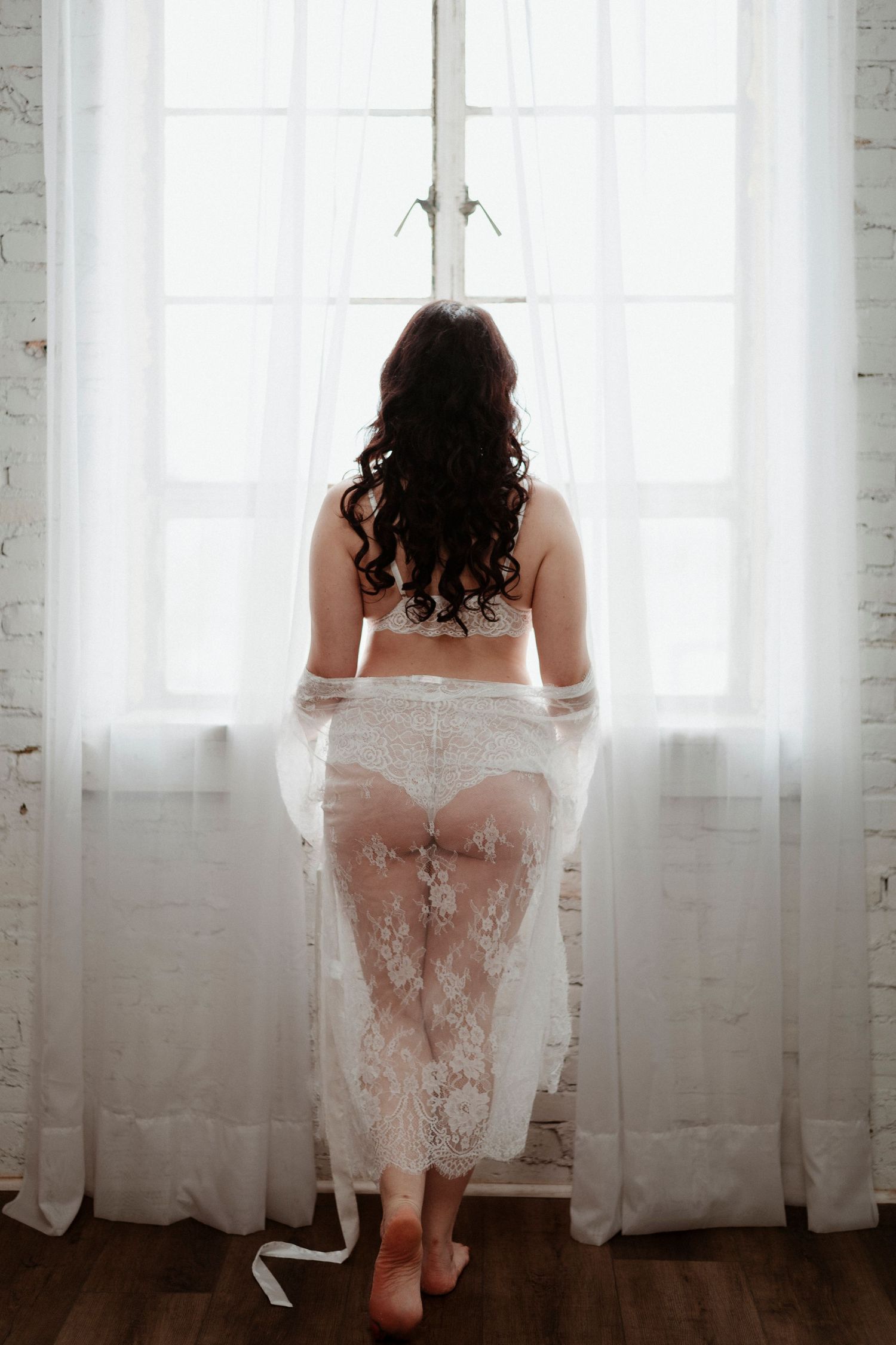 Back view of a figure in white lace standing by a bright window with sheer curtains and natural light.