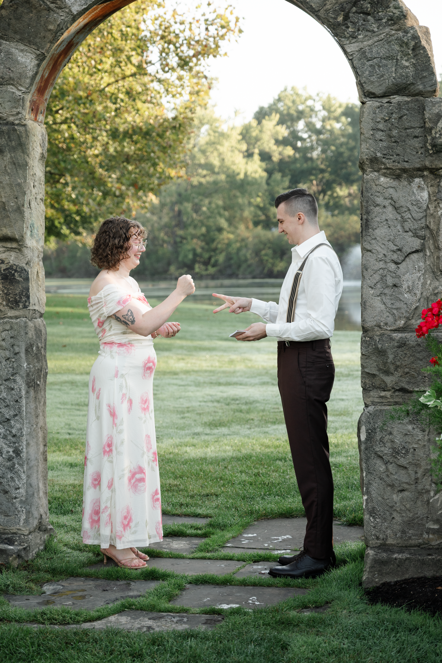 A couple stands under a stone archway in a garden setting, dressed in vintage-inspired attire while sharing a playful moment.