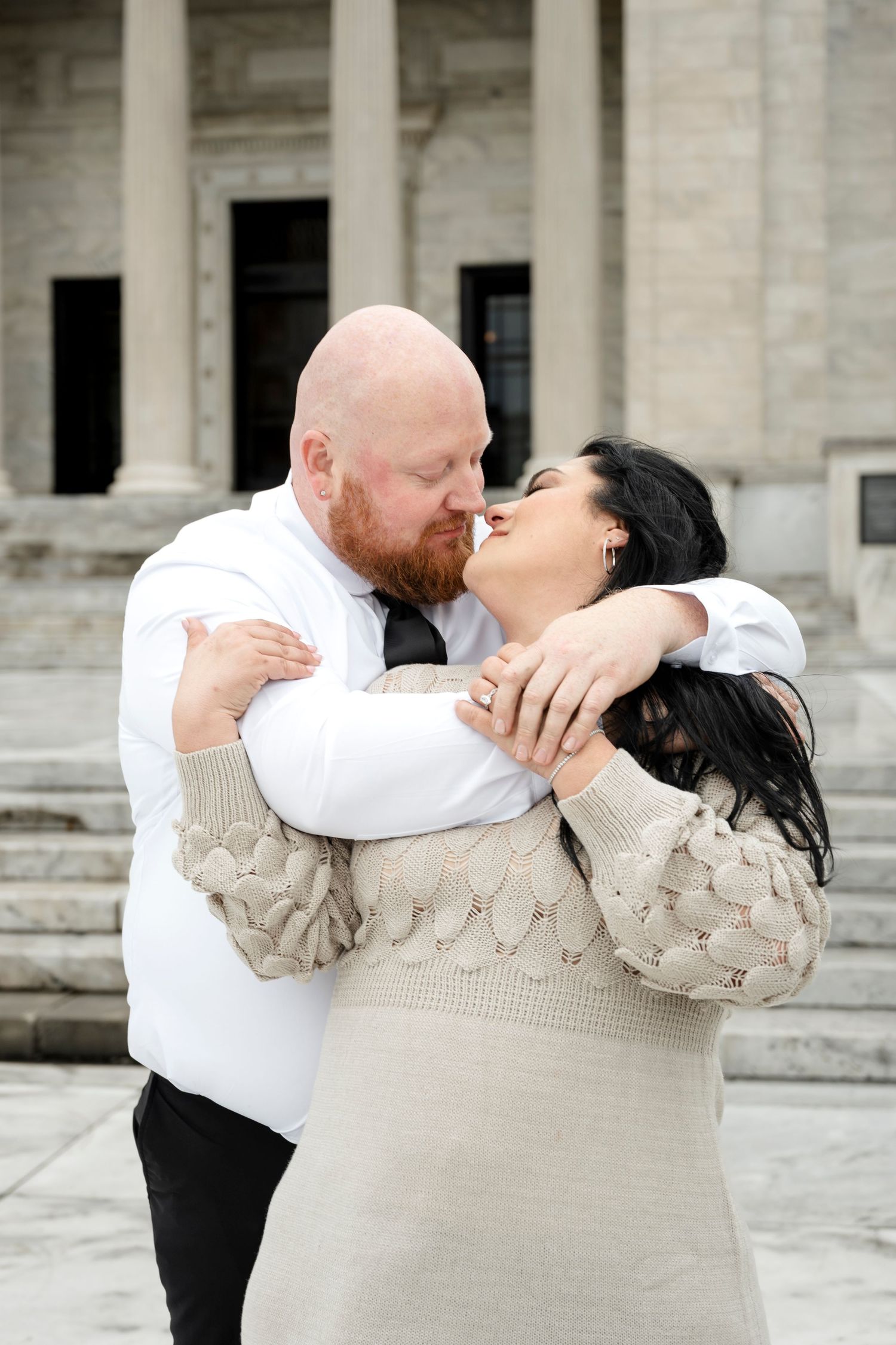 A bald man with a red beard kisses a dark-haired woman in a beige sweater dress outside a marble building.