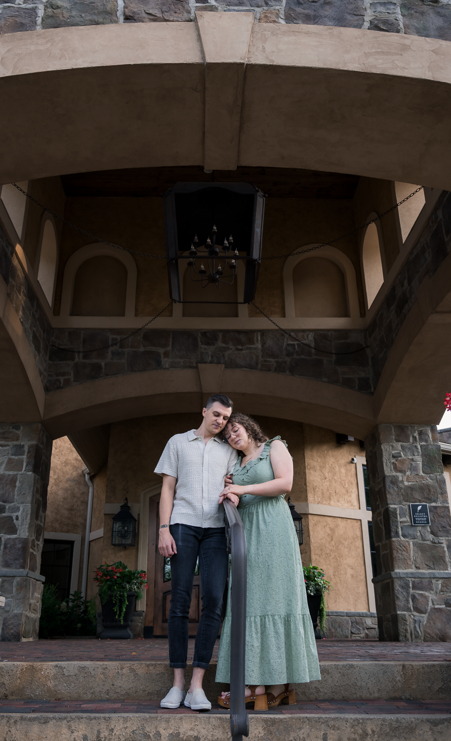 Couple posing romantically in front of a stone castle-style building, woman in green dress leaning on man's shoulder.