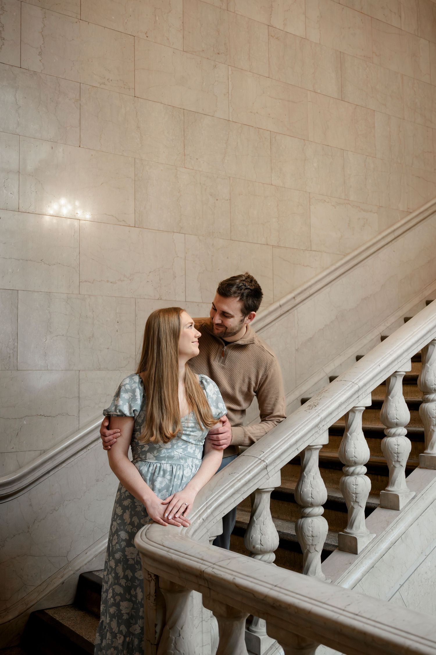 Couple sharing a romantic moment on elegant marble staircase, woman in floral dress, man in tan sweater.