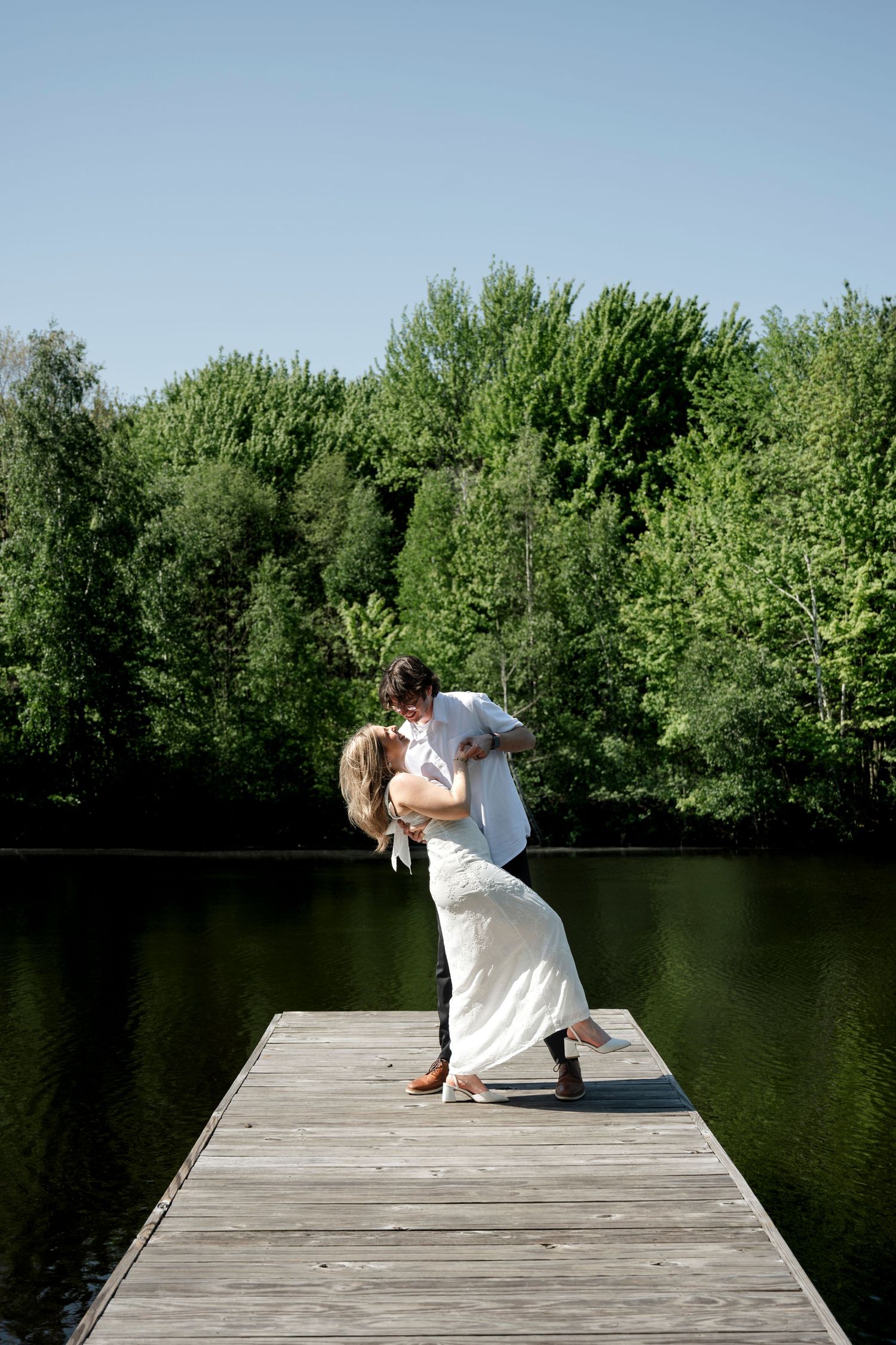 Couple sharing a romantic dip kiss on a wooden dock by a serene lake surrounded by lush green trees.