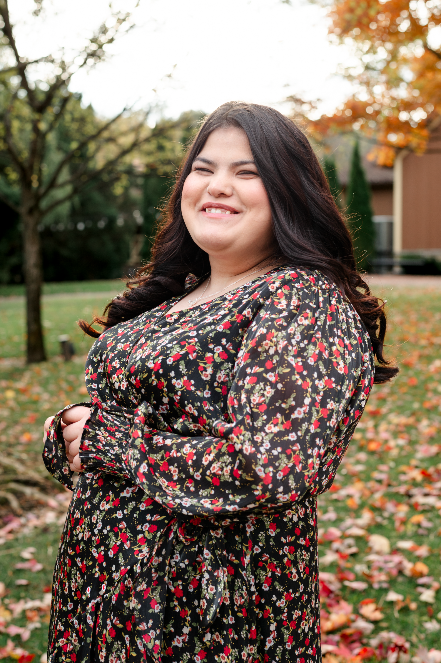 A person wearing a floral dress twirls playfully in a park with autumn leaves scattered on the ground.