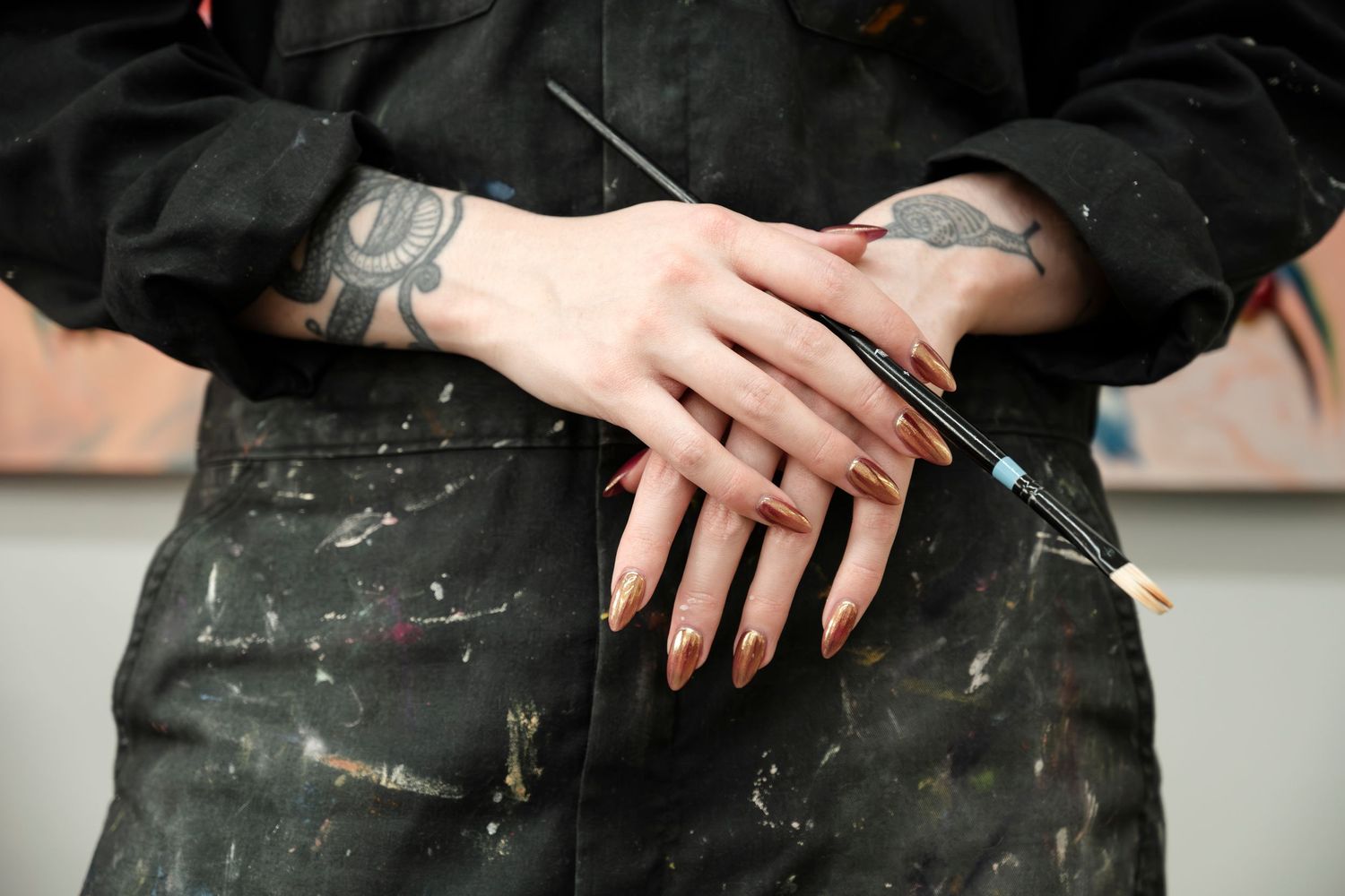 Paint-splattered hands with black nail polish rest on dark clothing, showing artistic wear and creative work.