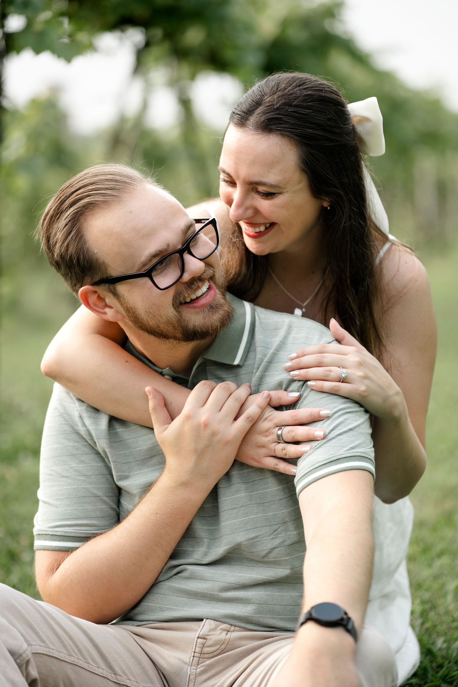 Happy couple embracing outdoors, man wearing glasses and woman with white bow smiling together in a green park setting.