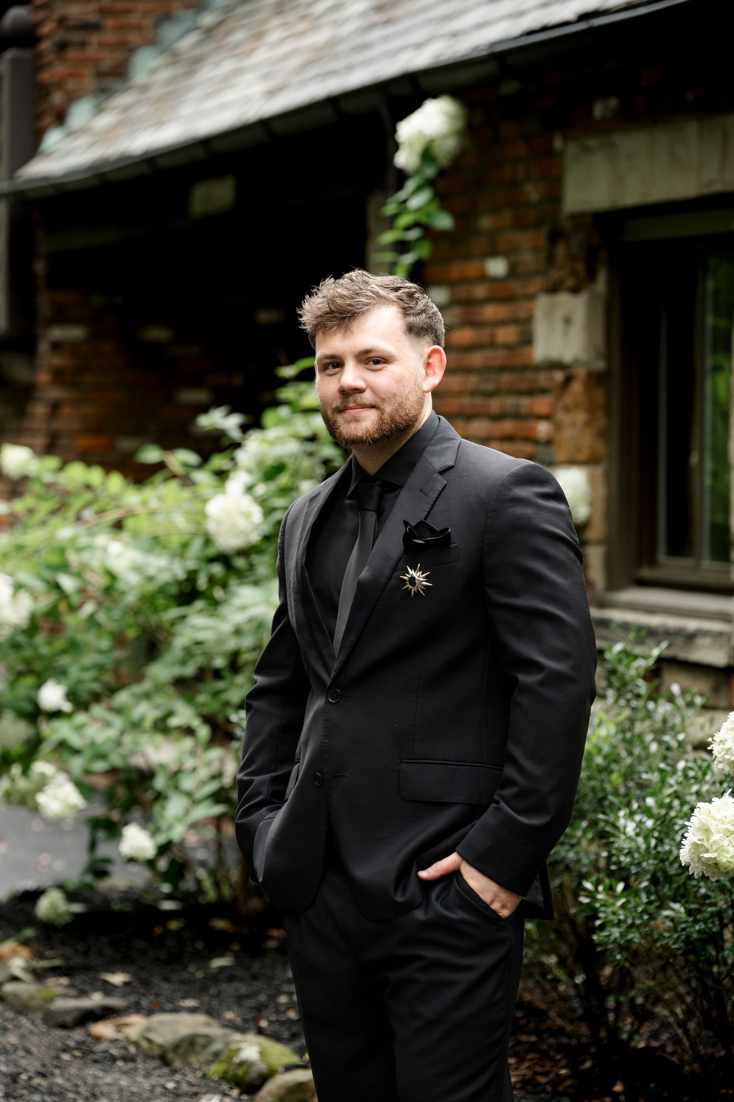 A groom in a black tuxedo with a star boutonniere poses outdoors near a brick building with white hydrangeas.