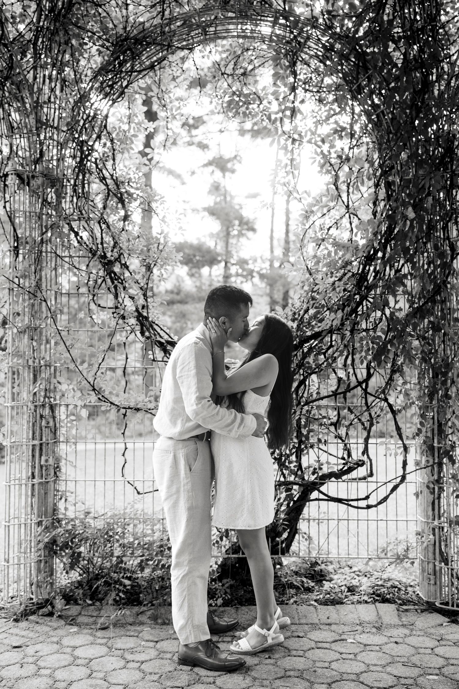 Black and white photo of a couple sharing a romantic kiss under a vine-covered garden arch.