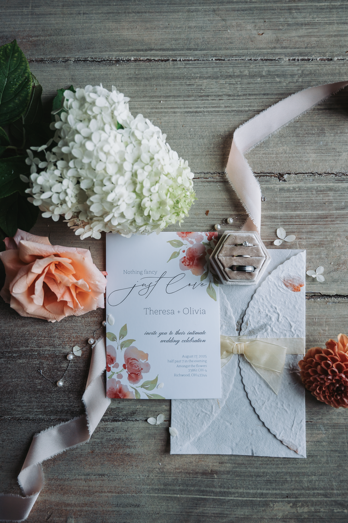 Wedding invitations with floral designs surrounded by coral roses and white hydrangeas on wooden surface.